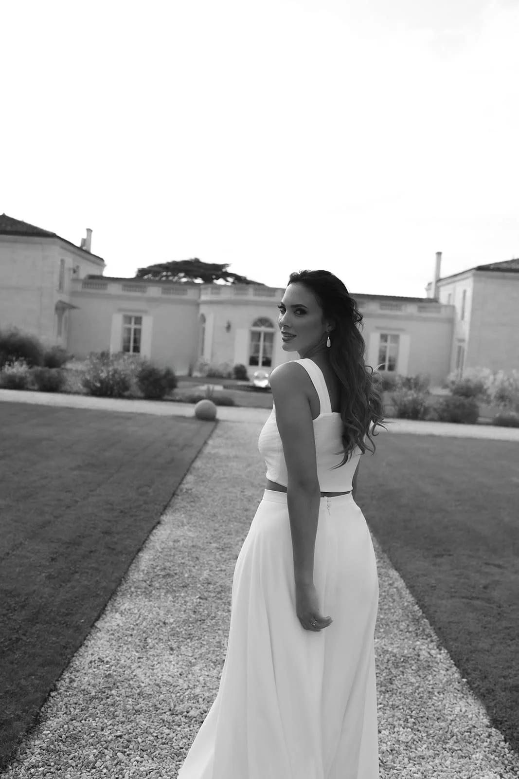 Une femme en robe blanche se tenant sur un chemin dans une propriété élégante, avec des bâtiments résidentiels en arrière-plan, en photo noire et blanc.
