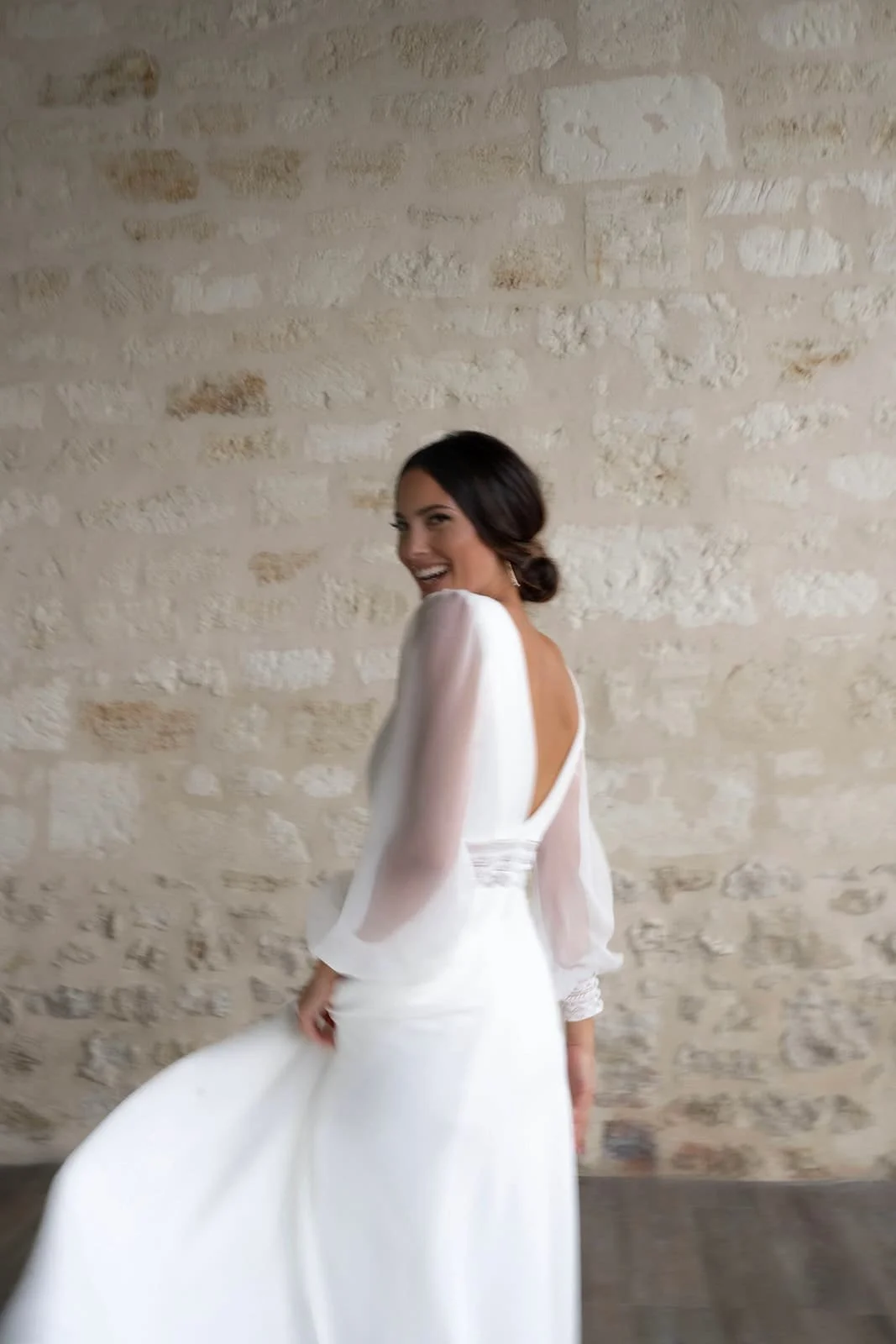 Femme en robe de mariée blanche souriant à l'appareil, debout contre un mur en pierre beige.