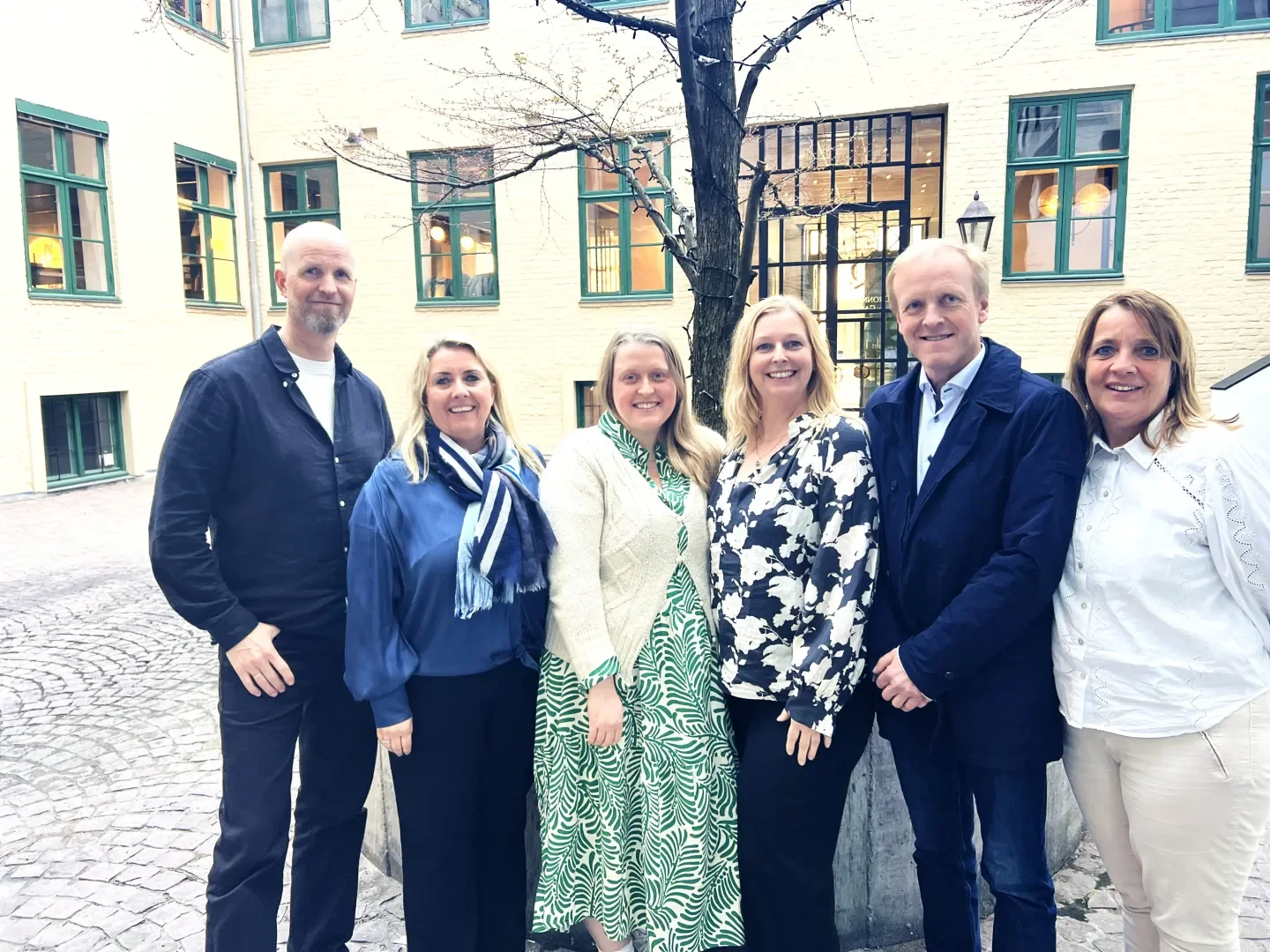 Syv personer står ute i en gårdsplass foran en gul vegg med grønne vinduer, smilende og poserer for foto.