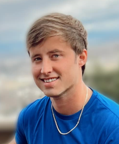 Young man with light brown hair smiling, wearing a blue shirt and a silver chain necklace outdoors.