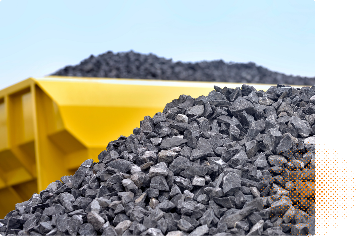 Close-up of gray gravel stones piled in front of a yellow container or dump truck, with a clear sky in the background.