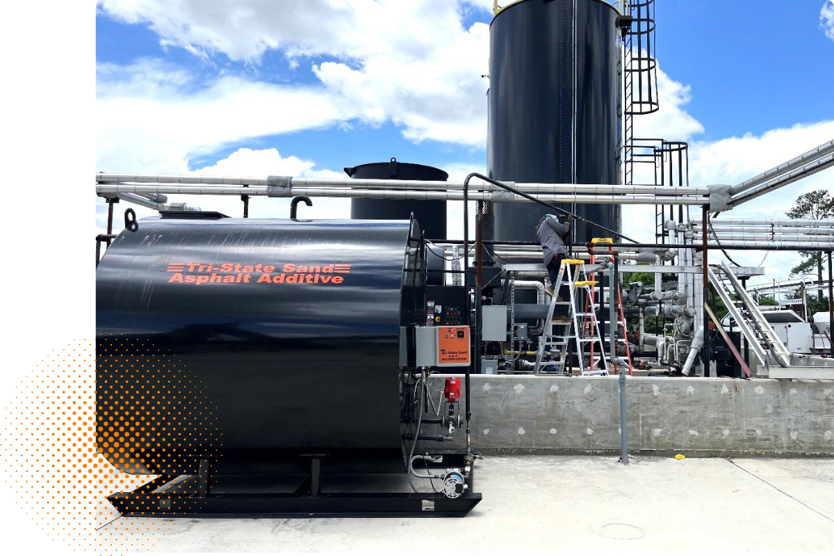 Industrial facility with a large black storage tank labeled 'Tri-state Sand Asphalt Additive' and various pipes, with a worker on a ladder working on equipment against a partly cloudy sky.
