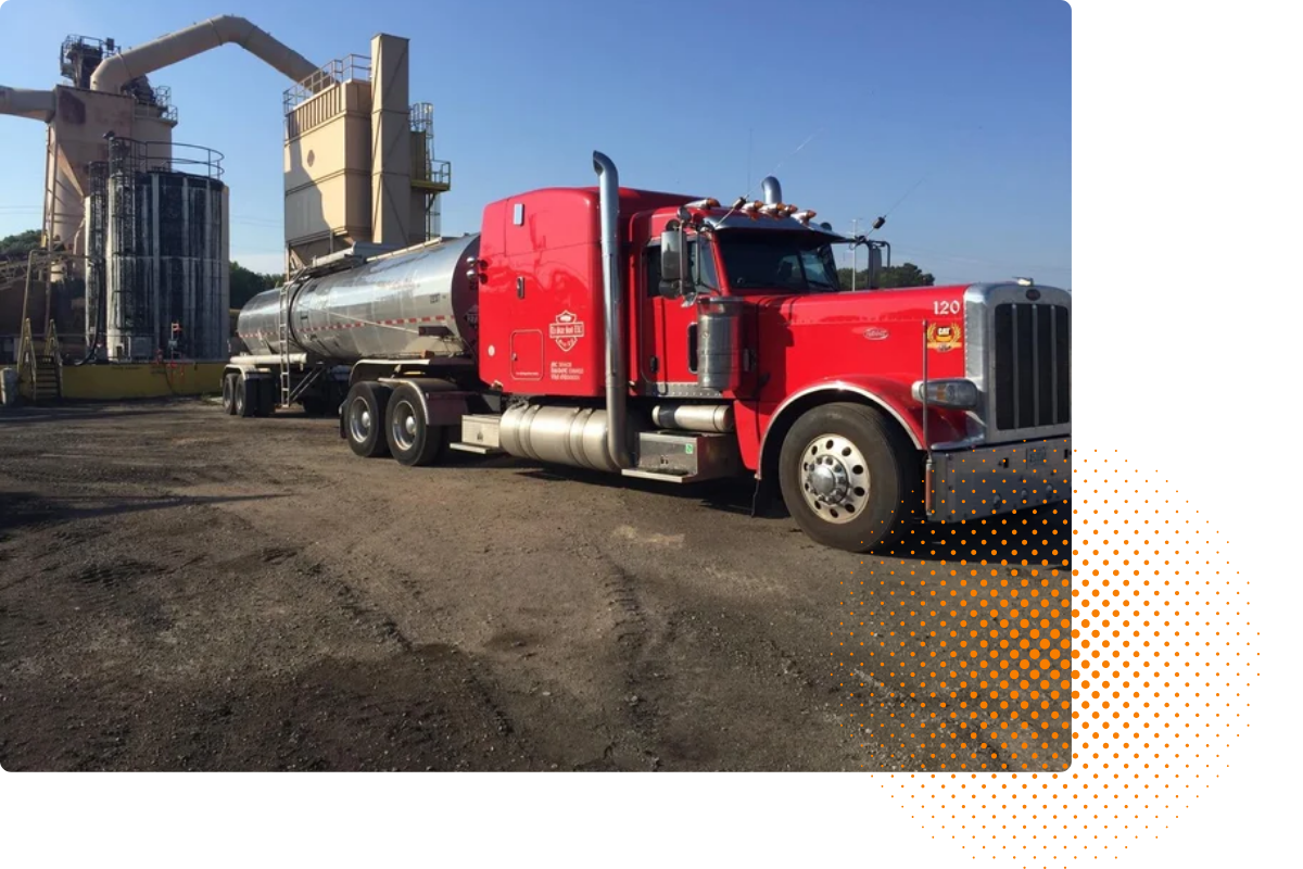 A large red tanker truck parked on a dirt lot near an industrial plant under a clear blue sky.