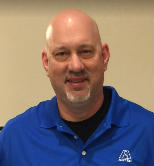 A bald man with a goatee wearing a blue collared shirt with the ASTEC logo, smiling at the camera.