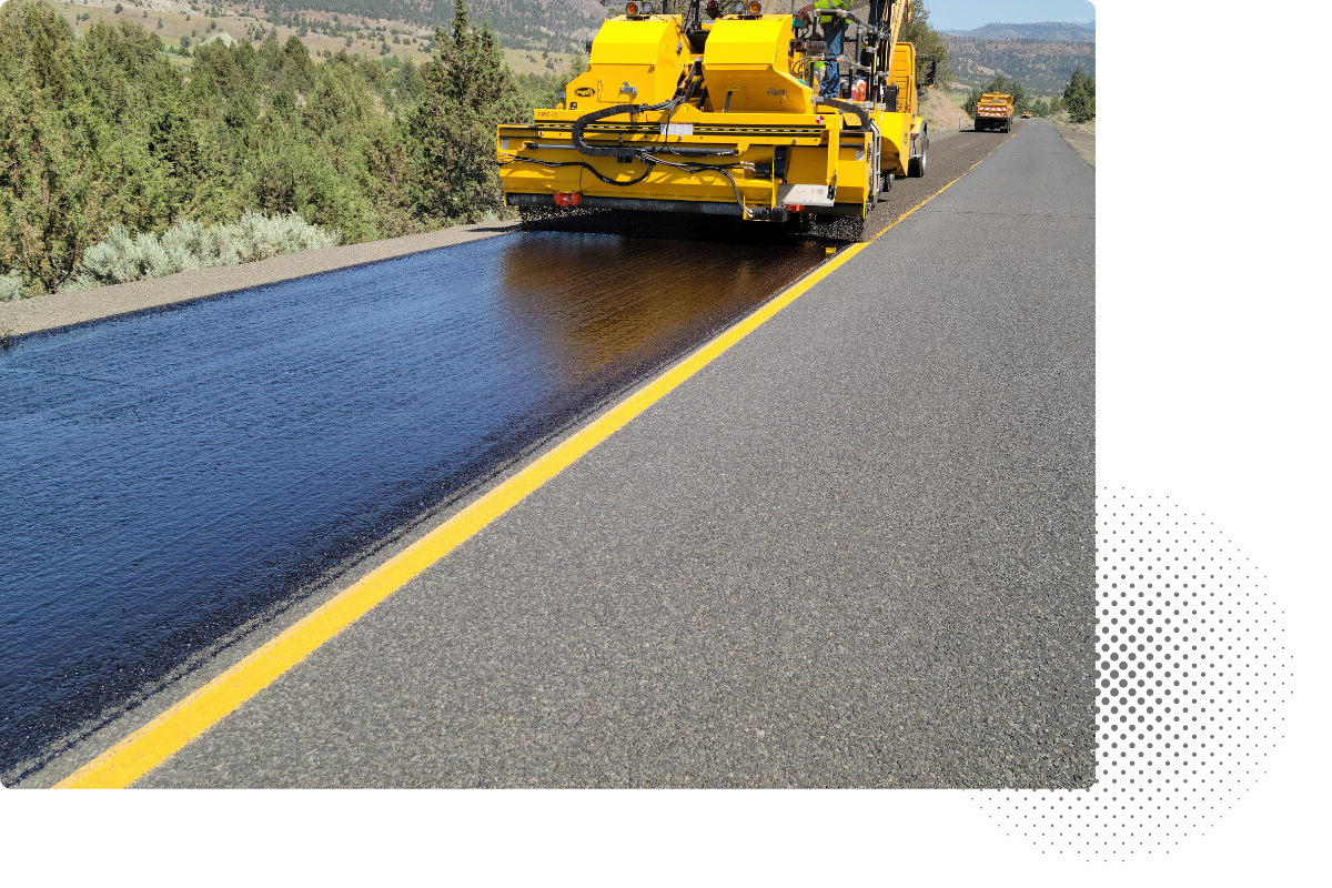 A road paving machine applies a fresh layer of asphalt onto the road surface, with a yellow line marking the edge of the lane, and trucks in the background.