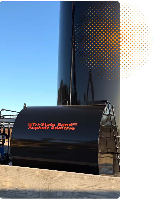 Close-up of a black industrial tank or container with the label "Tri-State Sand Asphalt Additive" in red, set outdoors against a clear blue sky, with a large black structure and some metal framework in the background.
