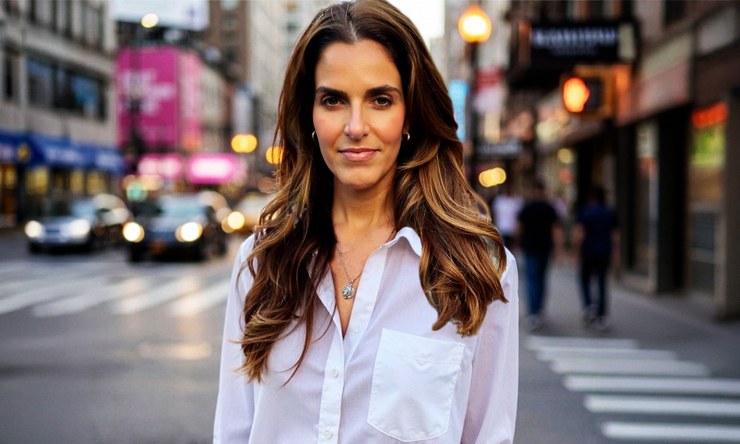 A woman with long wavy brown hair wearing a white shirt and hoop earrings standing on a busy city street at dusk.