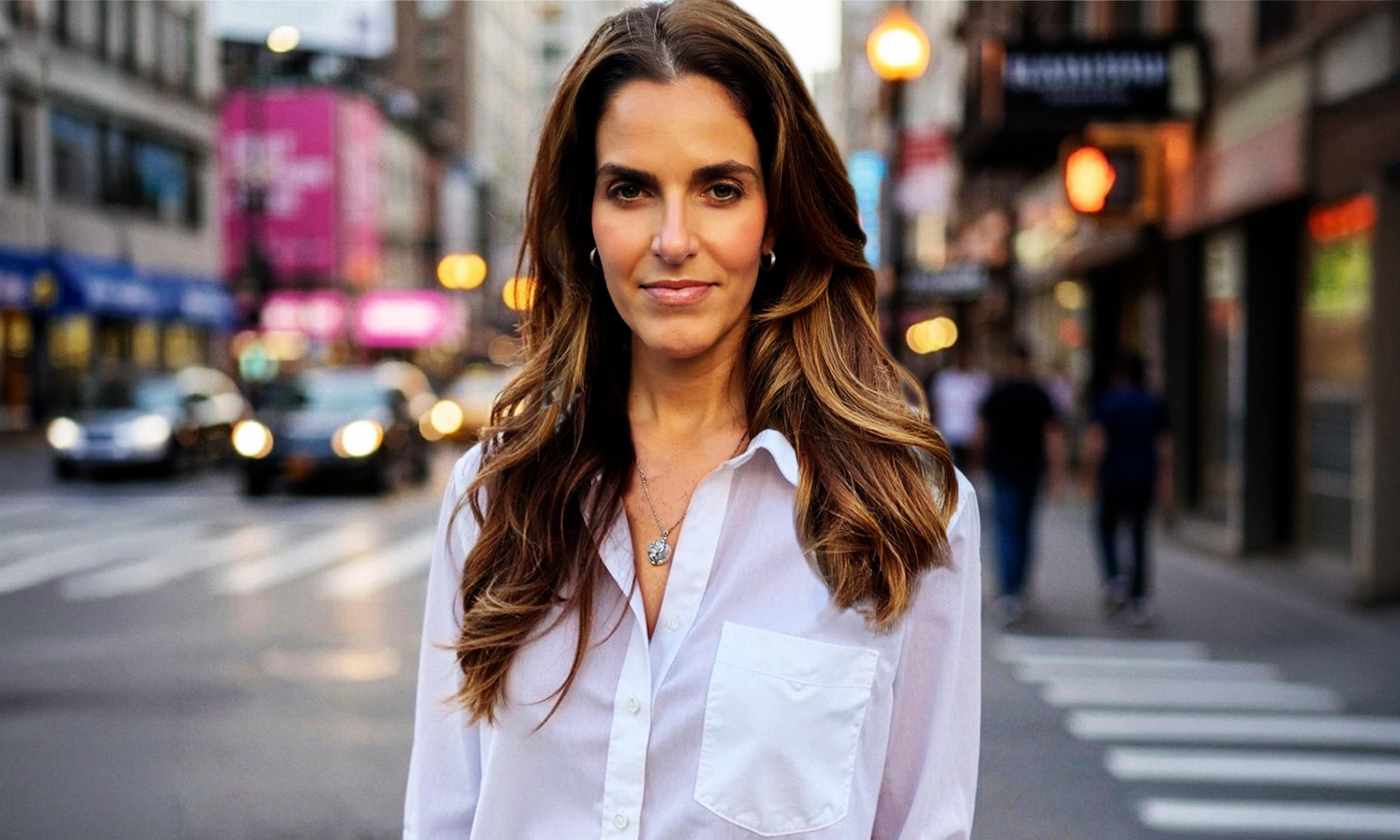 A woman with long wavy brown hair wearing a white shirt and hoop earrings standing on a busy city street at dusk.