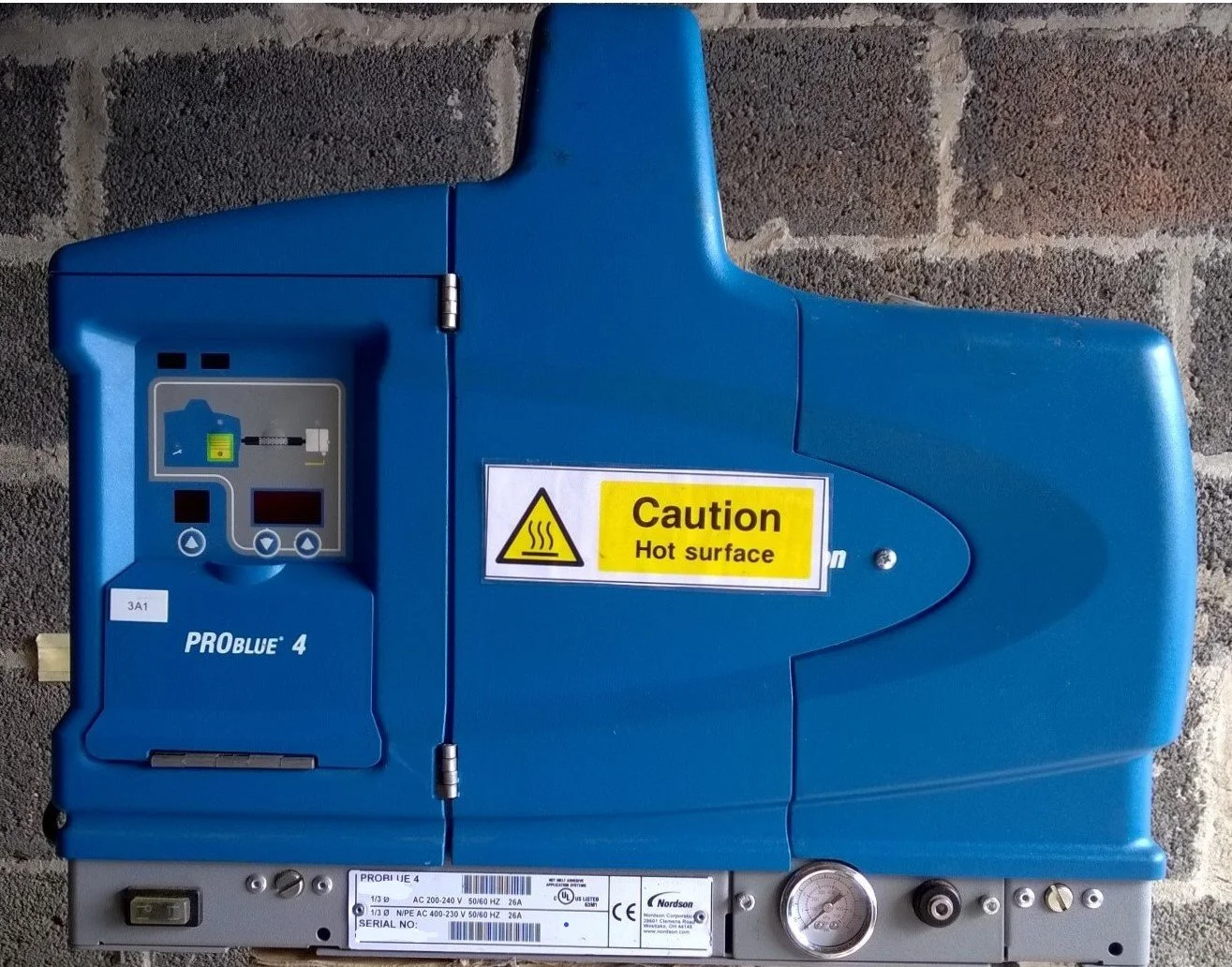 Blue industrial machine with a warning label caution hot surface, showing digital controls, pressure gauge, and power connections, set against a brick wall background.