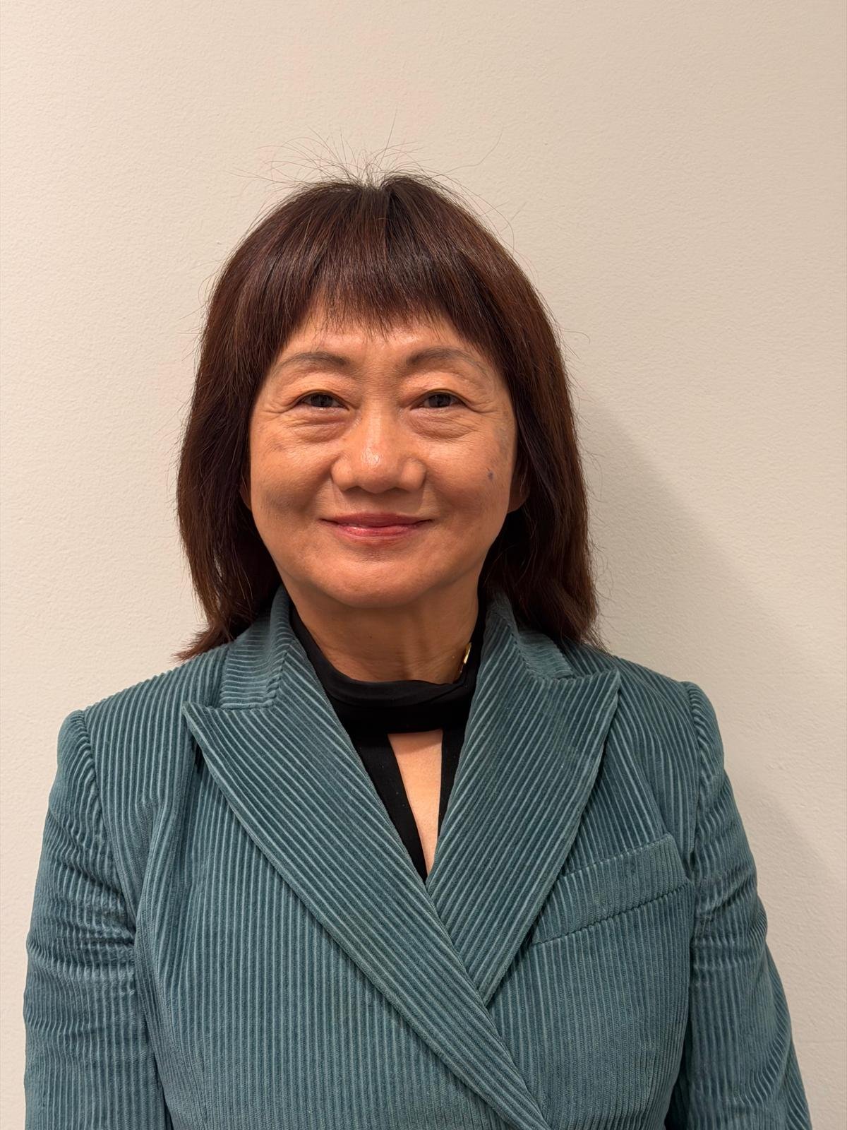 A woman with shoulder-length brown hair and a friendly expression, wearing a teal pinstripe blazer and a black top, standing against a plain light-colored wall.
