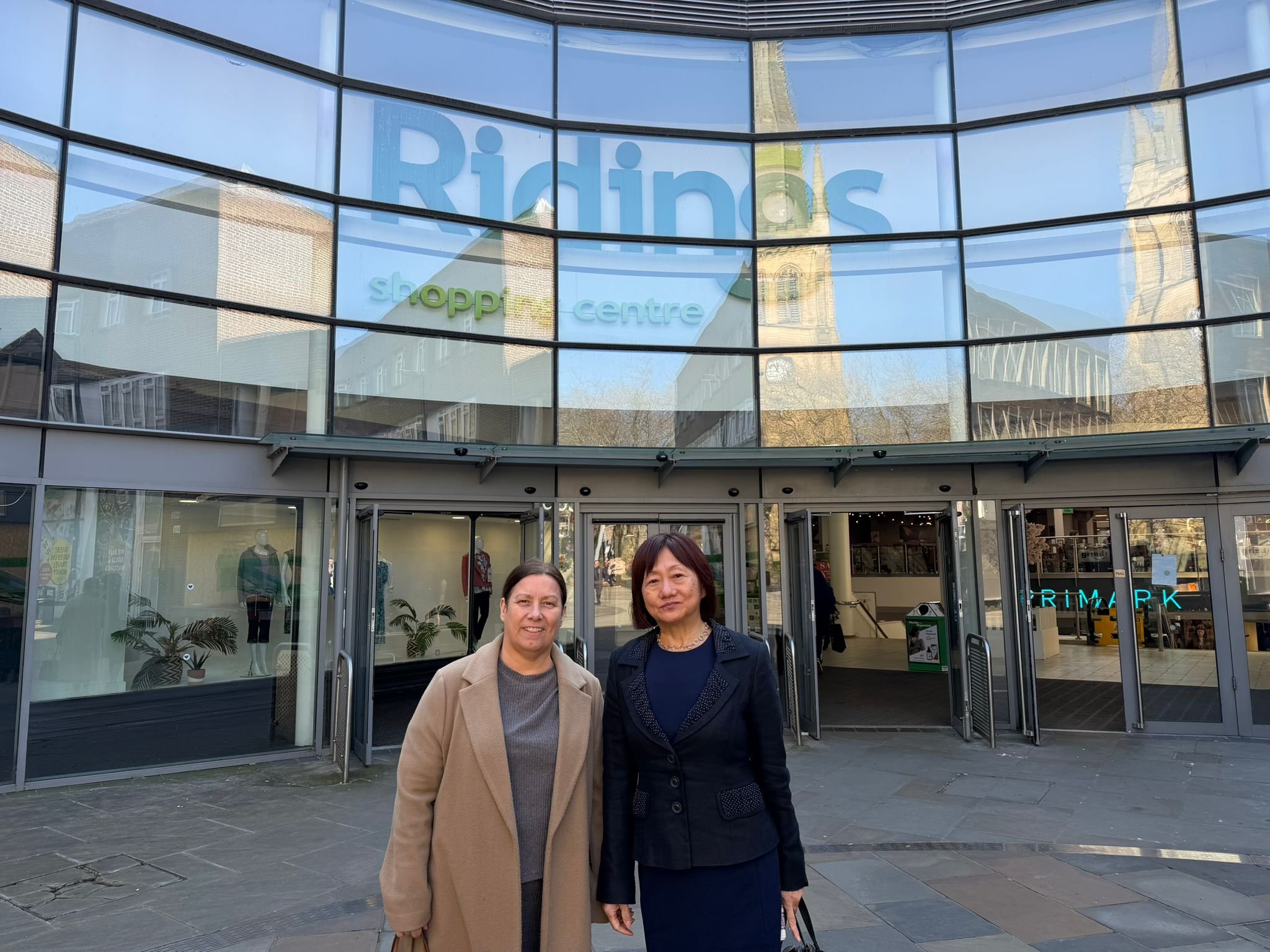 Two women standing in front of a modern glass shopping center entrance, with reflections of nearby buildings and a church spire visible in the glass. The shopping center has large signs, including one that says 'Ridings'.