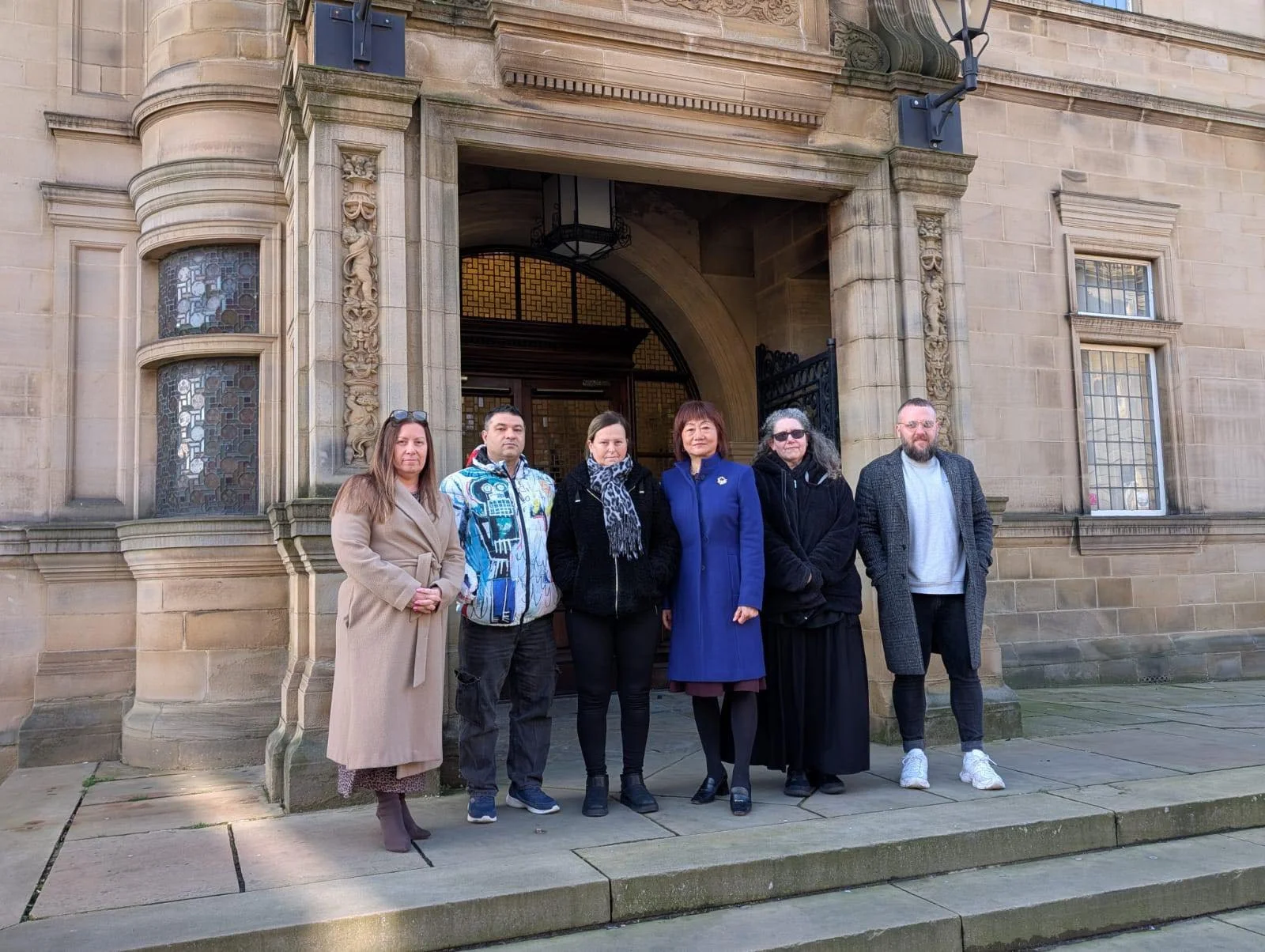 Six people standing on steps outside an ornate stone building with arched doorway and decorative windows.