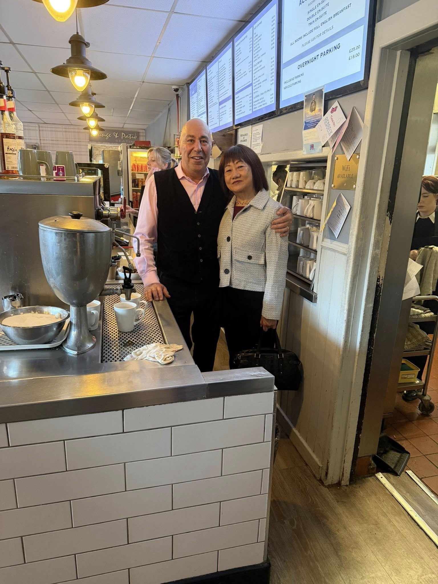A man and a woman standing together at a cafe counter, smiling at the camera, with menu screens above them and coffee-making equipment in the foreground.