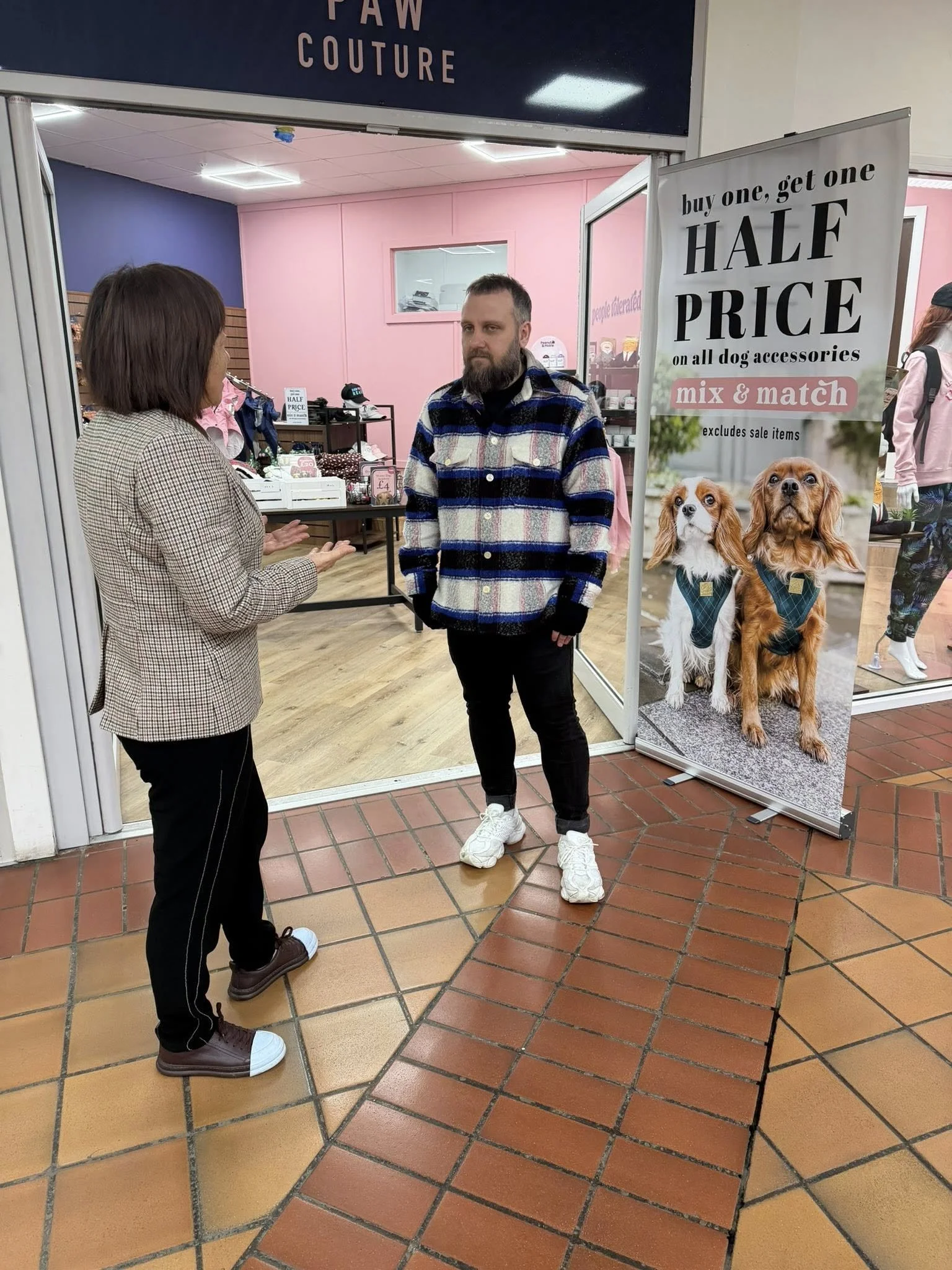 A man and woman are having a conversation outside a pet store called 'Paw Couture.' The man has a beard and is wearing a plaid jacket and white sneakers. The woman has shoulder-length hair and is wearing a checkered blazer and dark pants. There is a 