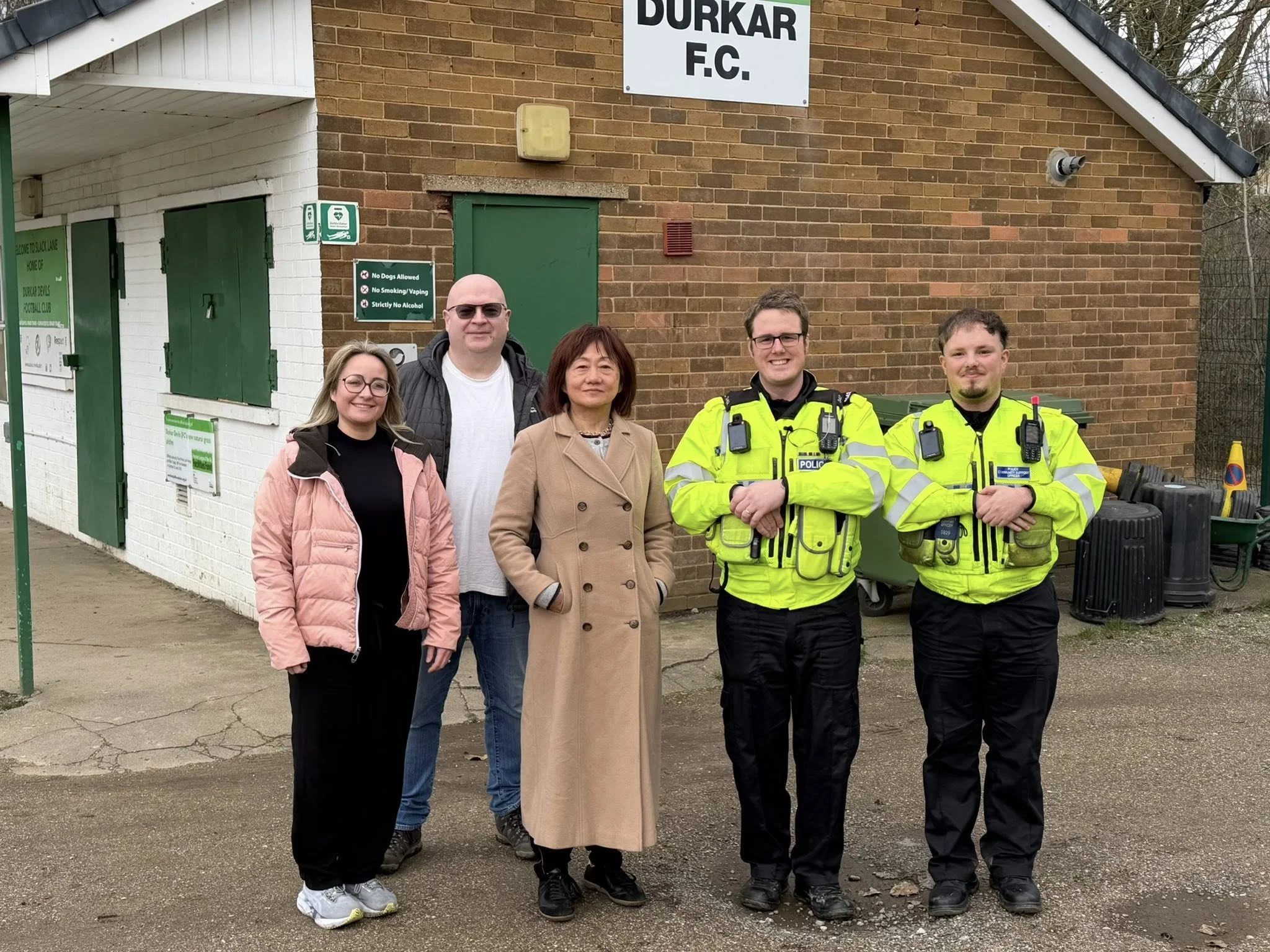 Group of five people standing outside, including two police officers in neon yellow jackets, two women, and one man, in front of a brick building with a sign reading 'DURKAR F.C.'