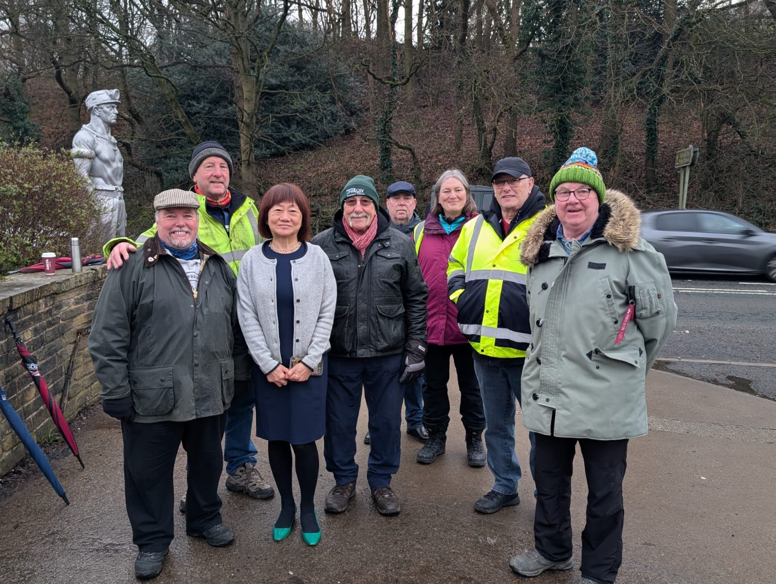 A group of eight people standing outdoors on a cloudy day, dressed in warm clothing, with trees and a statue of a soldier in the background, some wearing high visibility jackets.