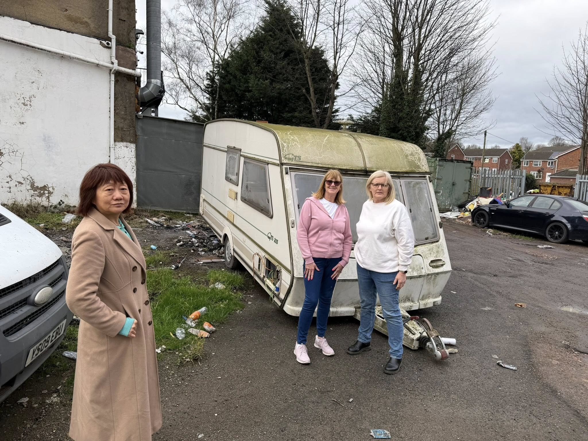 Three women standing in a parking lot near an abandoned, dirty caravan with moss and dirt on its roof, surrounded by trash and debris.