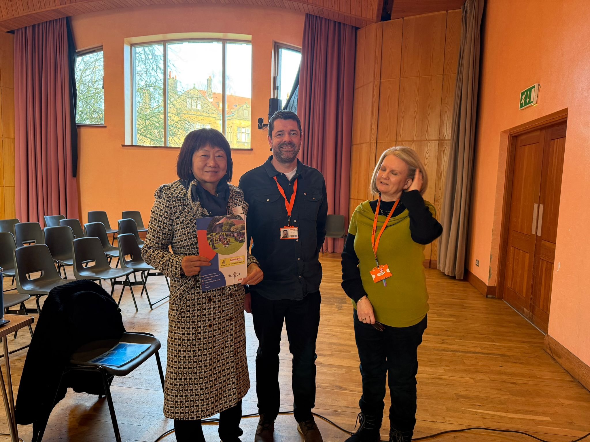 Three people standing in a room with wooden floors, large windows, pink curtains, and orange walls. The woman on the left holds a promotional flyer, the man in the middle, and the woman on the right are smiling. Chairs are arranged in the background,