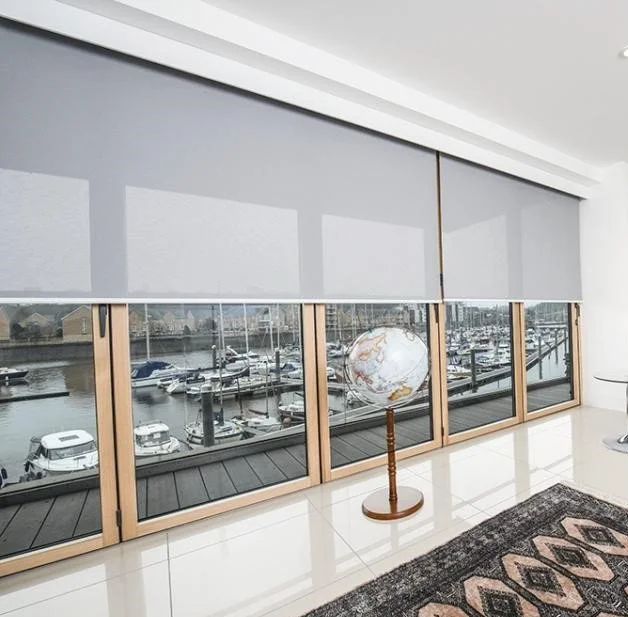 Interior room with large windows overlooking marina with boats, a globe on a stand, roller shades partially lowered, and a patterned rug on tile floor.