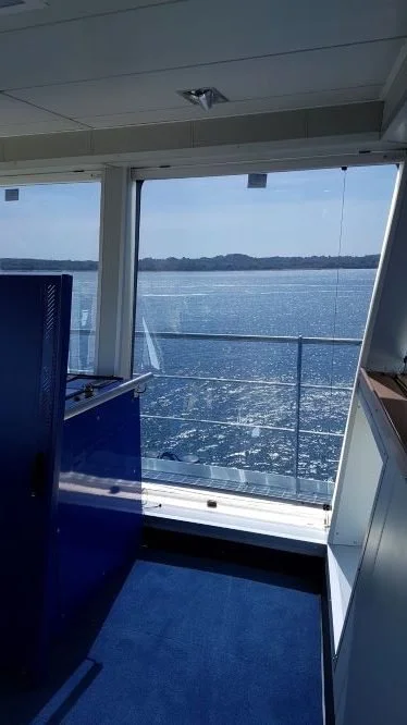View from inside a boat's cabin showing a large window overlooking water and distant land with hills, and part of the boat's interior including a blue counter and windows.