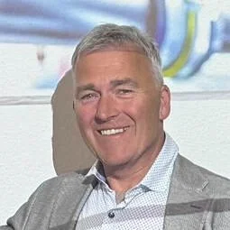 A middle-aged man with short gray hair, wearing a light gray suit jacket and a light blue dress shirt, smiling in an indoor setting.
