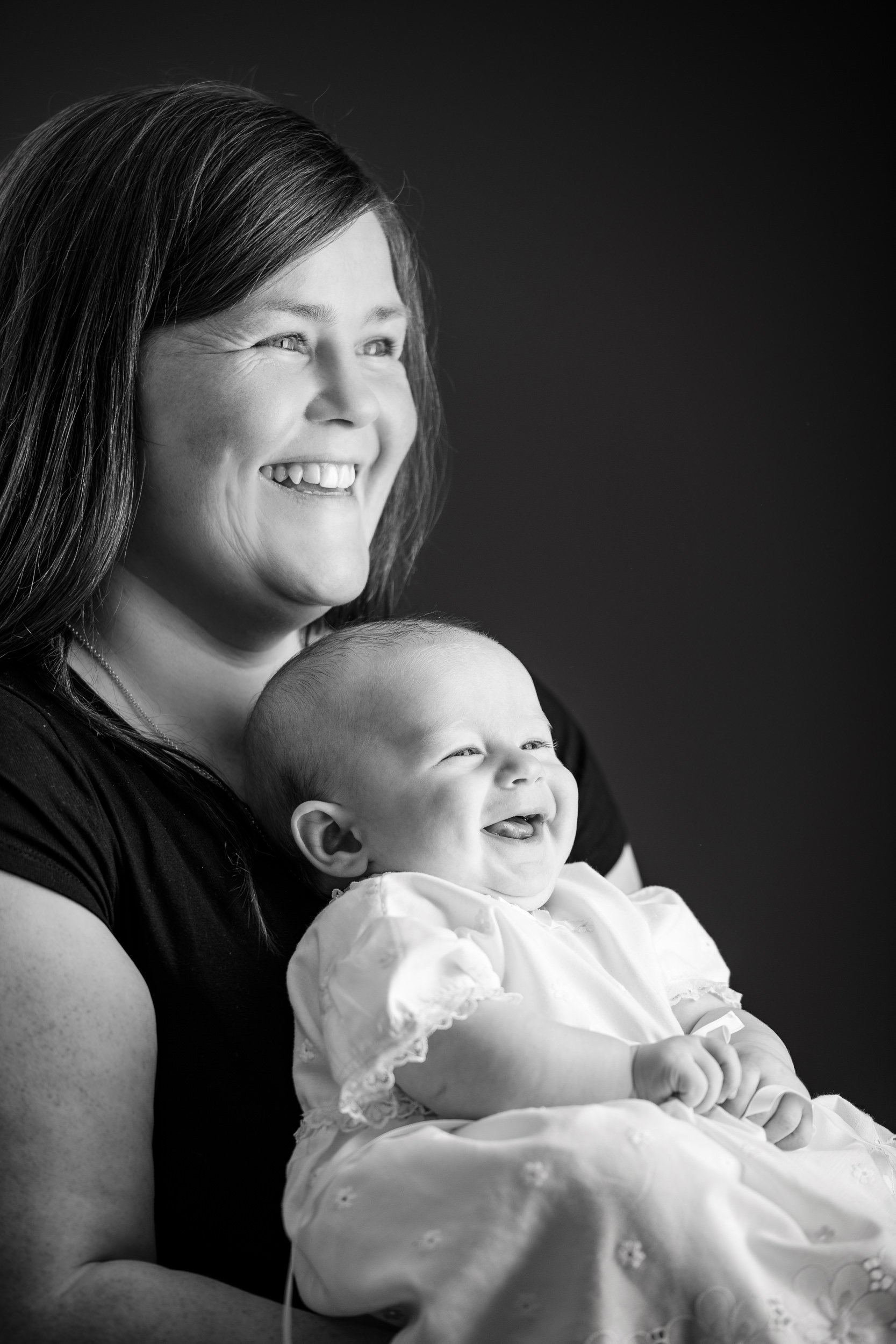 A woman holding a smiling baby girl on her lap, both joyful in a black and white portrait.
