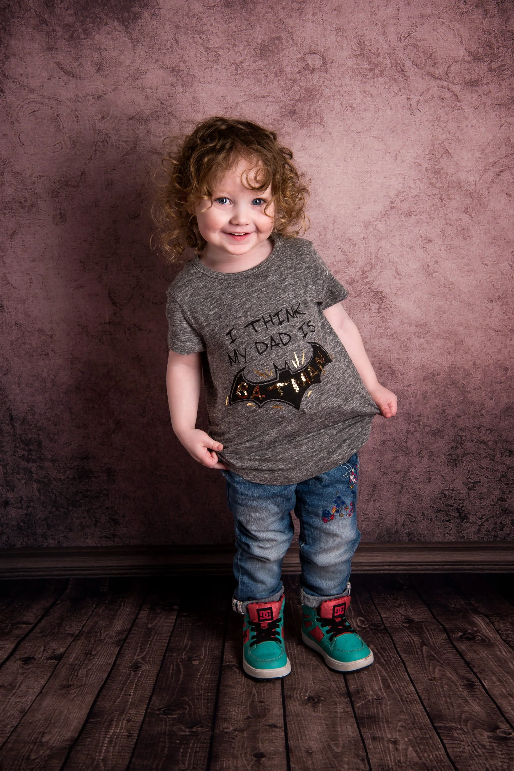 A young girl with curly hair standing on a wooden floor in front of a textured pink wall. She is wearing a gray T-shirt with the text 'I think my dad is' and a bat graphic, along with faded blue jeans and teal sneakers with pink and black accents.