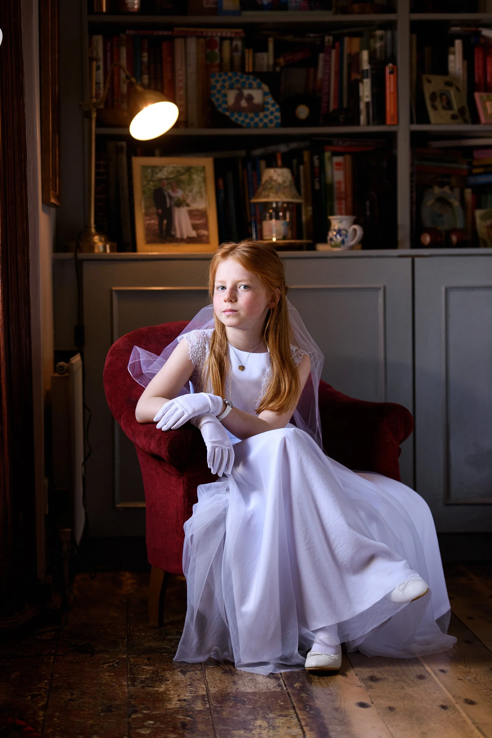 A young girl with red hair in a white dress and veil sitting on a red velvet chair in a cozy room with bookshelves, framed photos, and a lamp.