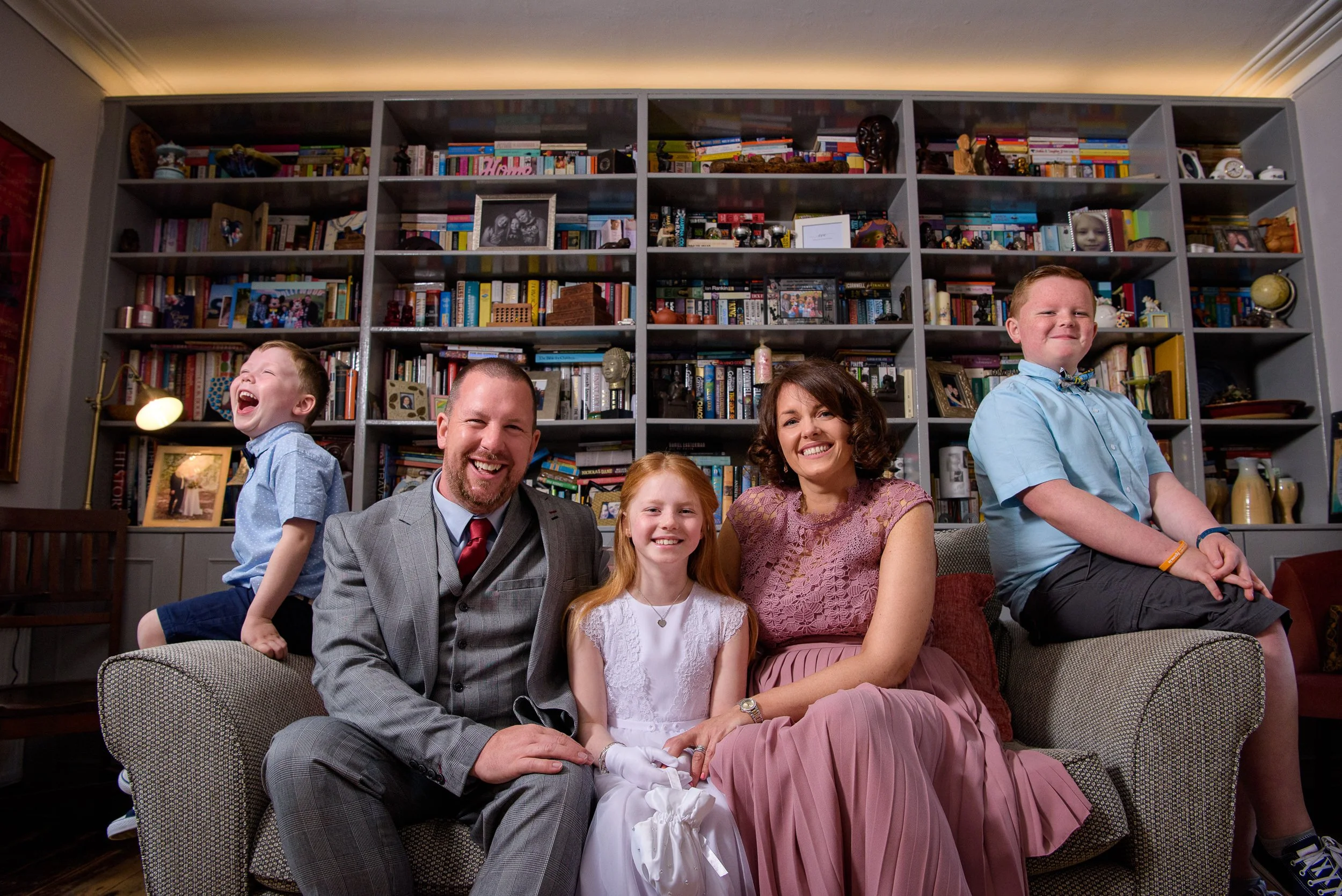 A family of five sitting on a sofa in a living room, smiling and laughing, with a large bookshelf filled with books and decorative items behind them.