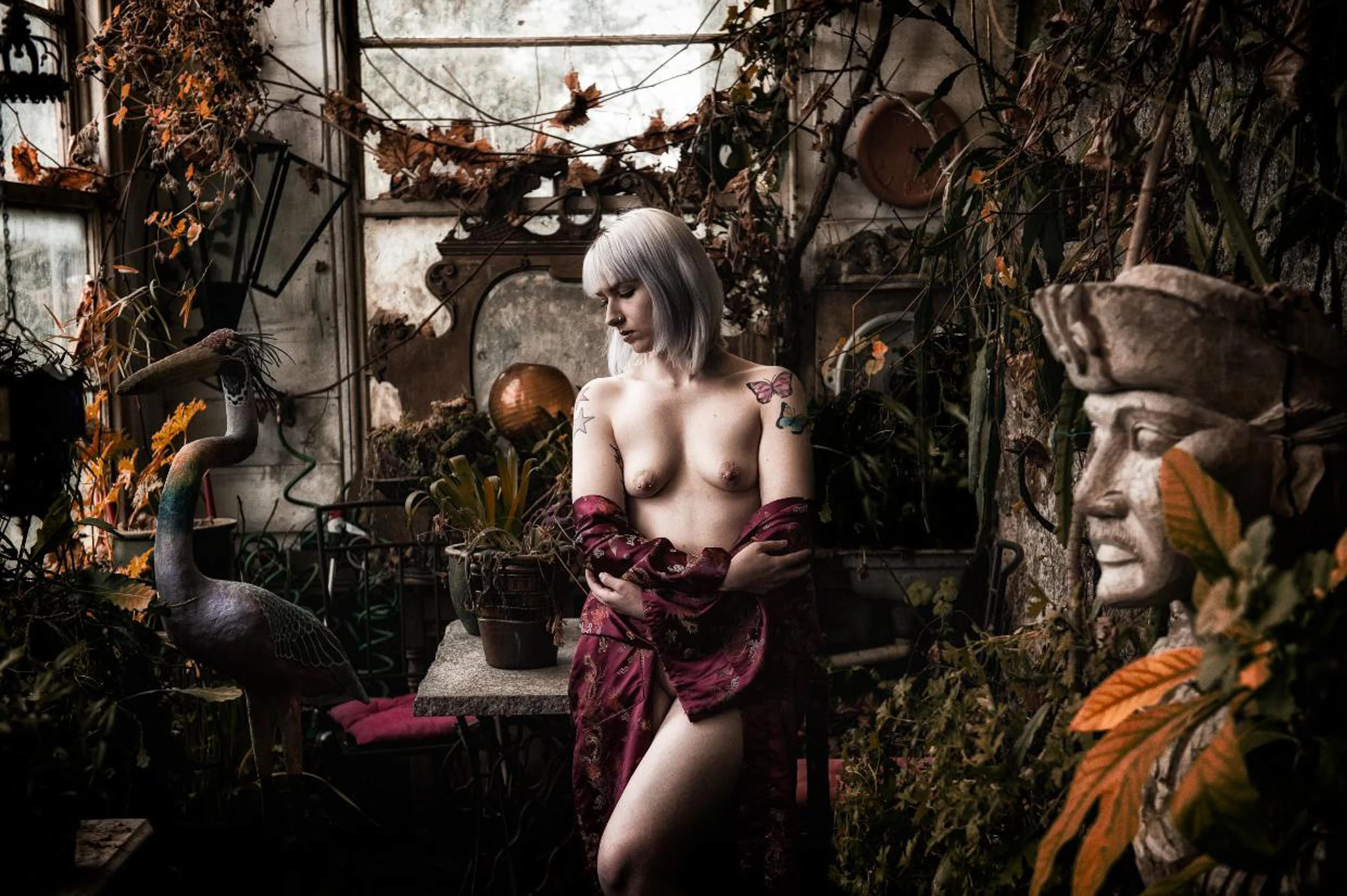 A woman with short grey hair and tattoos standing in a lush greenhouse, partially draped in fabric, surrounded by plants and vintage décor.
