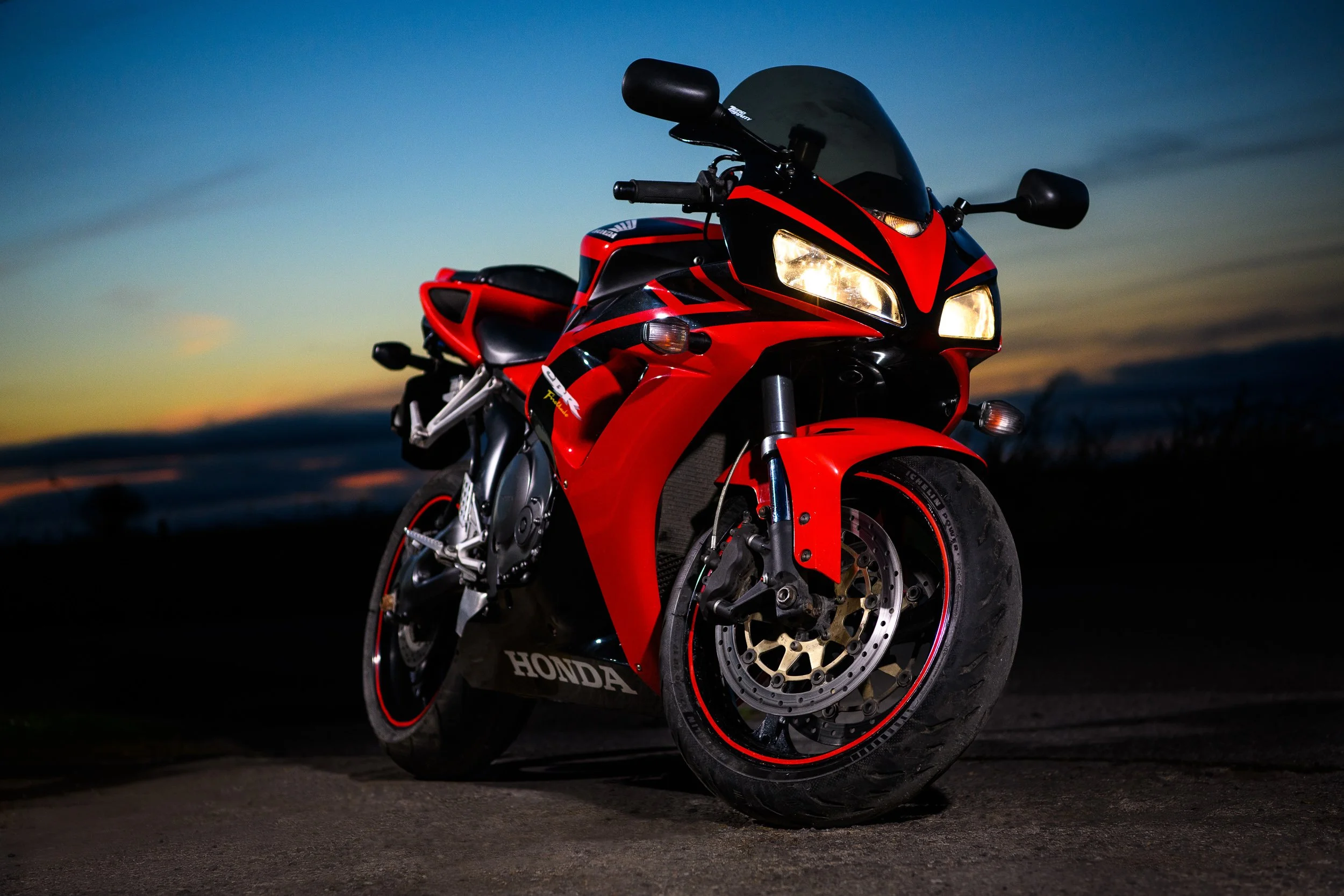 Honda sport motorcycle in red and black with headlights on, photographed on a dark road at sunset.
