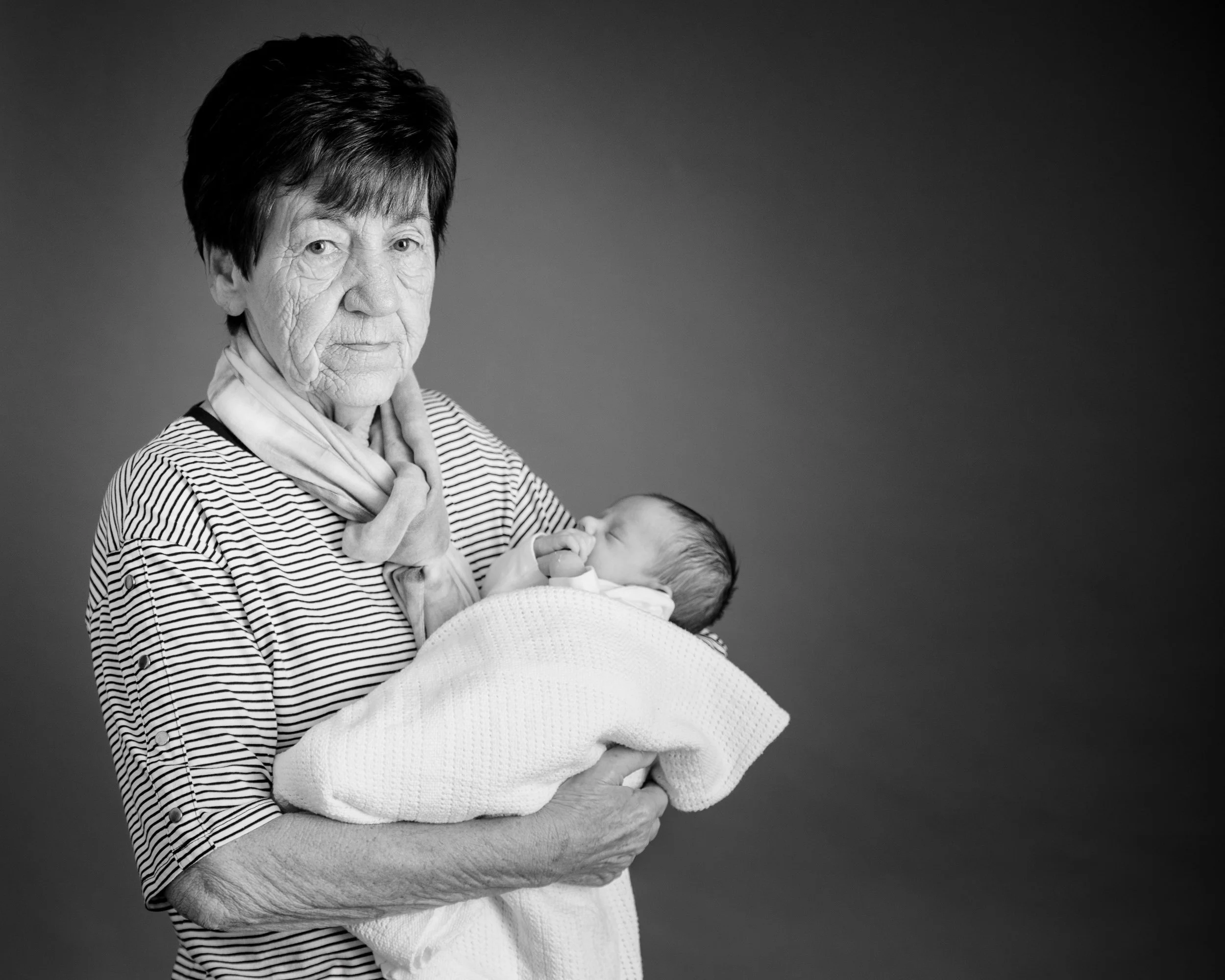An elderly woman holding a newborn baby wrapped in a white blanket against a soft grey background.
