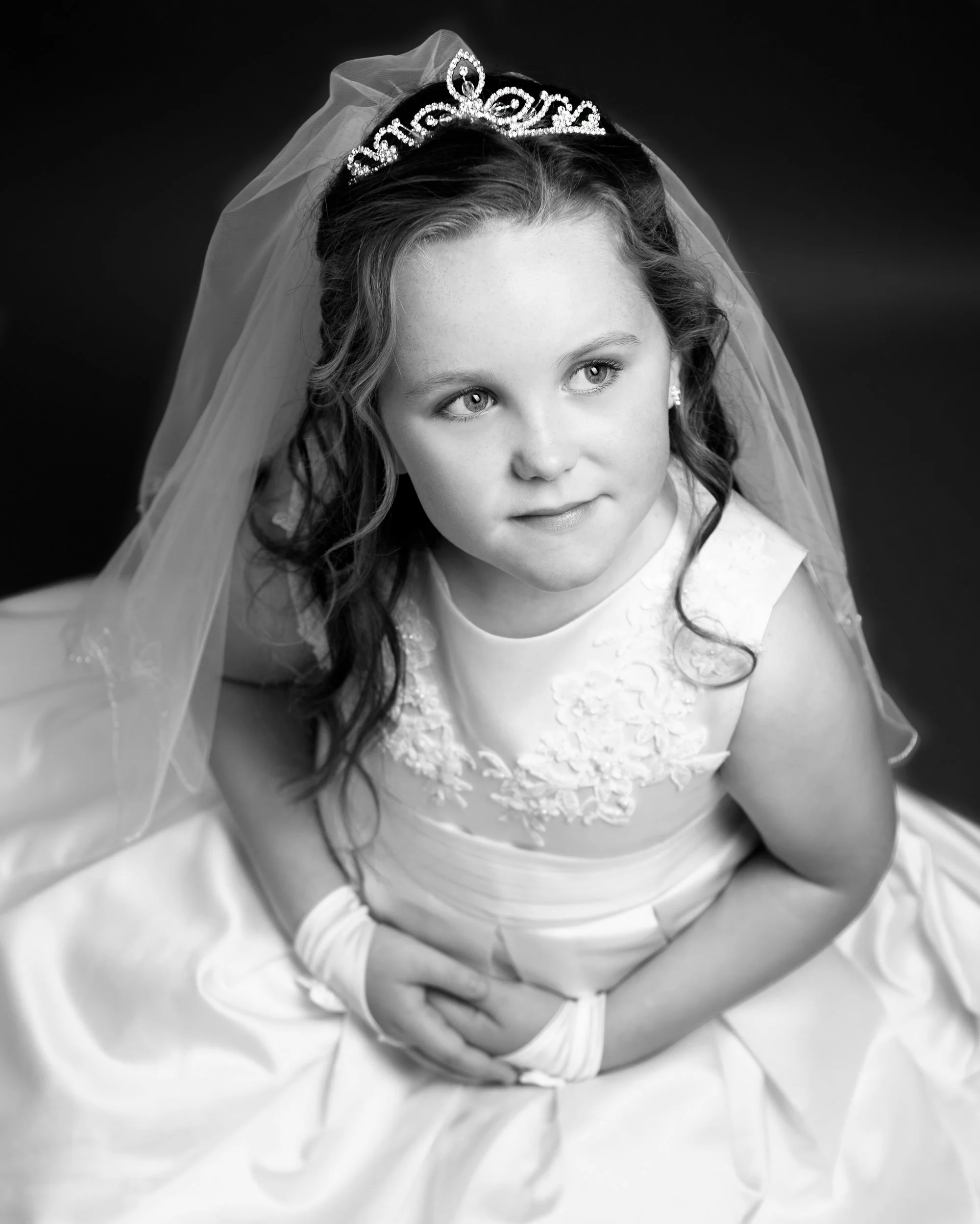 A young girl in a wedding dress and veil, wearing a tiara, looking up with a gentle expression.