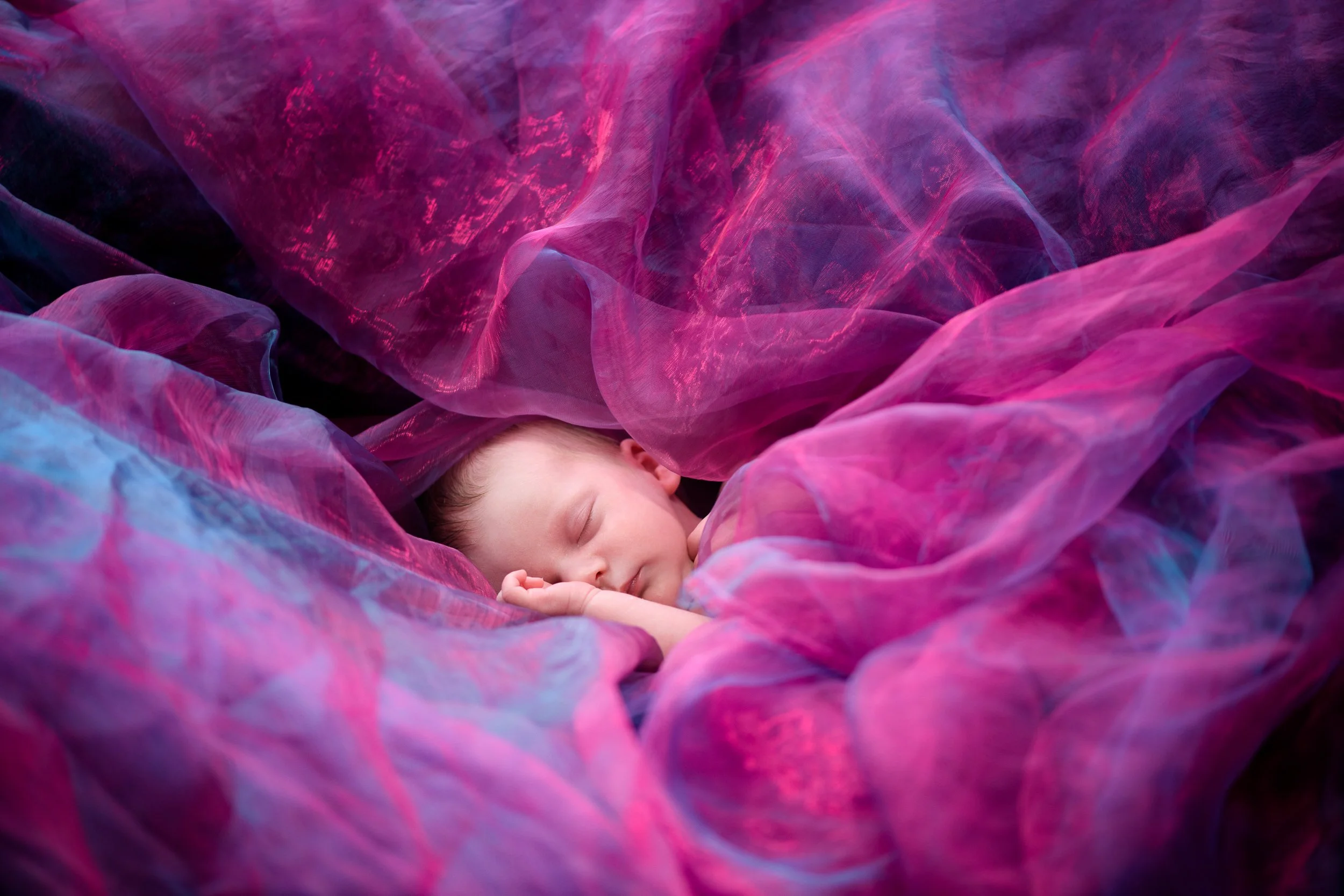 A newborn baby peacefully sleeping surrounded by soft pink and purple fabric.
