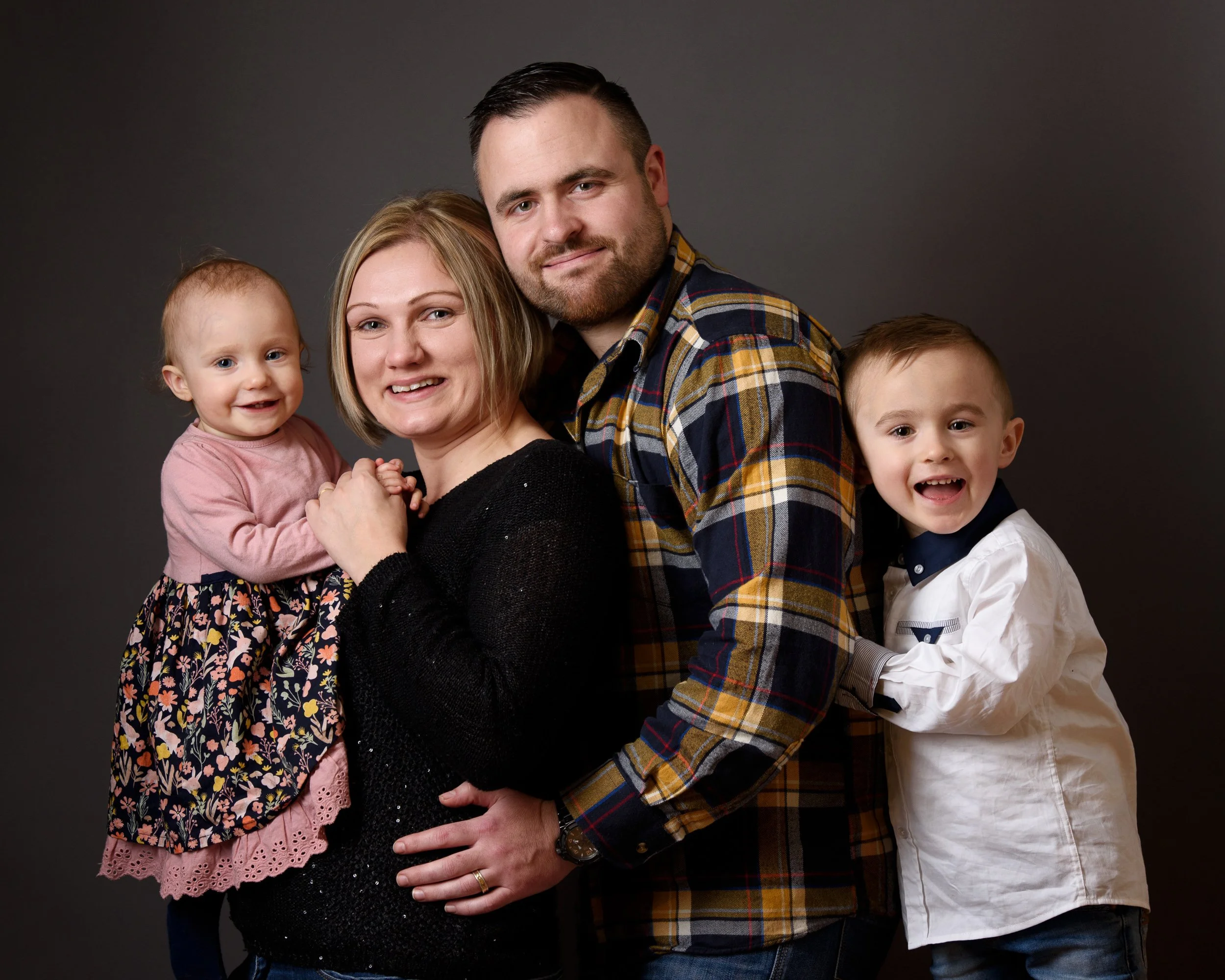 Family of four smiling and embracing together against a dark grey background.
