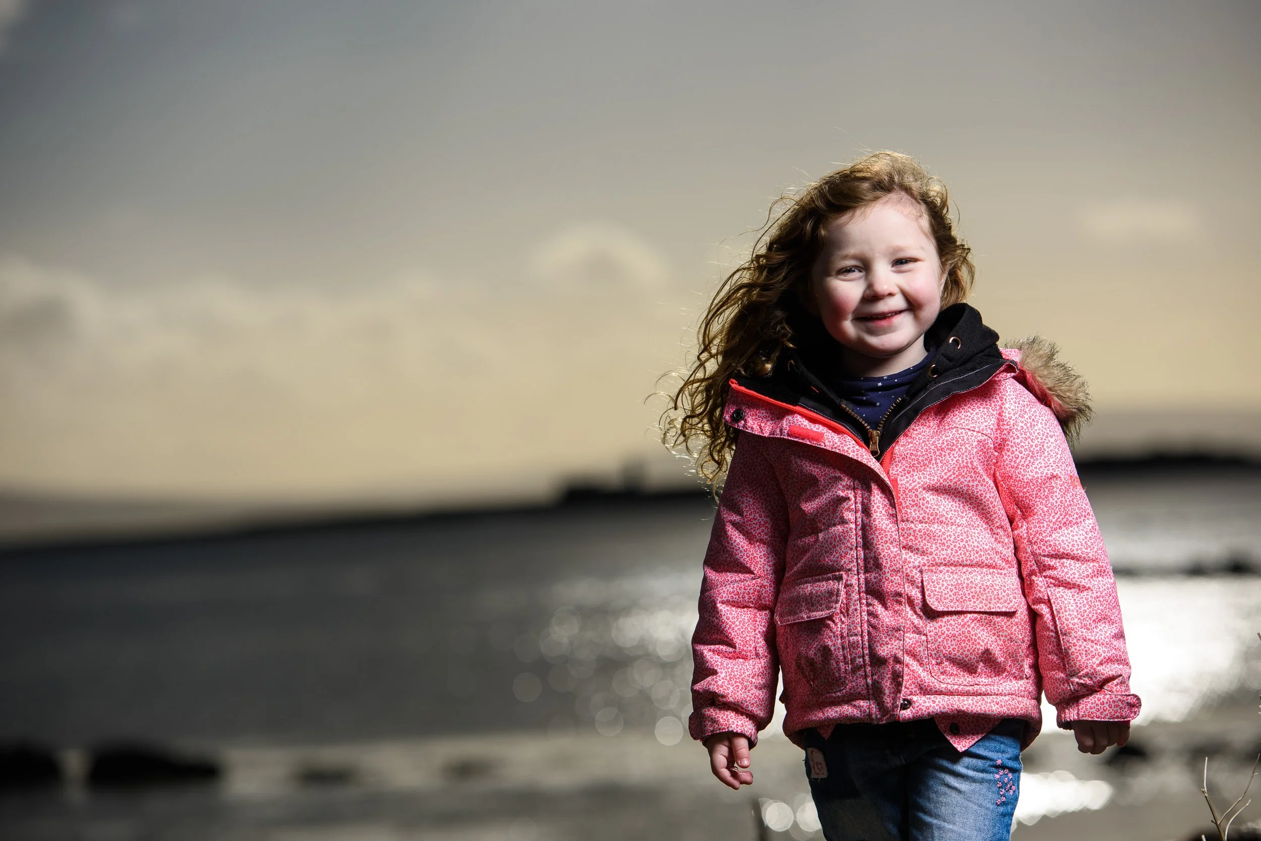 Young girl with curly hair smiling outdoors in front of a body of water at sunset or sunrise.