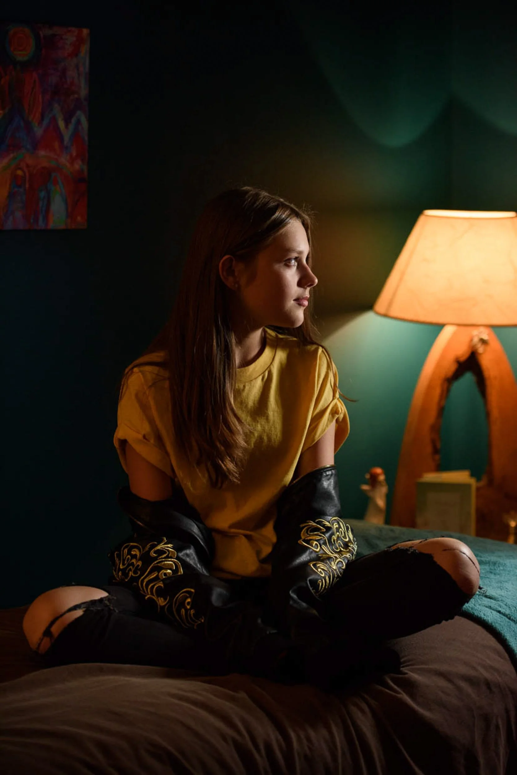 Moody indoor portrait of a woman sitting on a bed in warm lamplight wearing a yellow shirt and gloves
