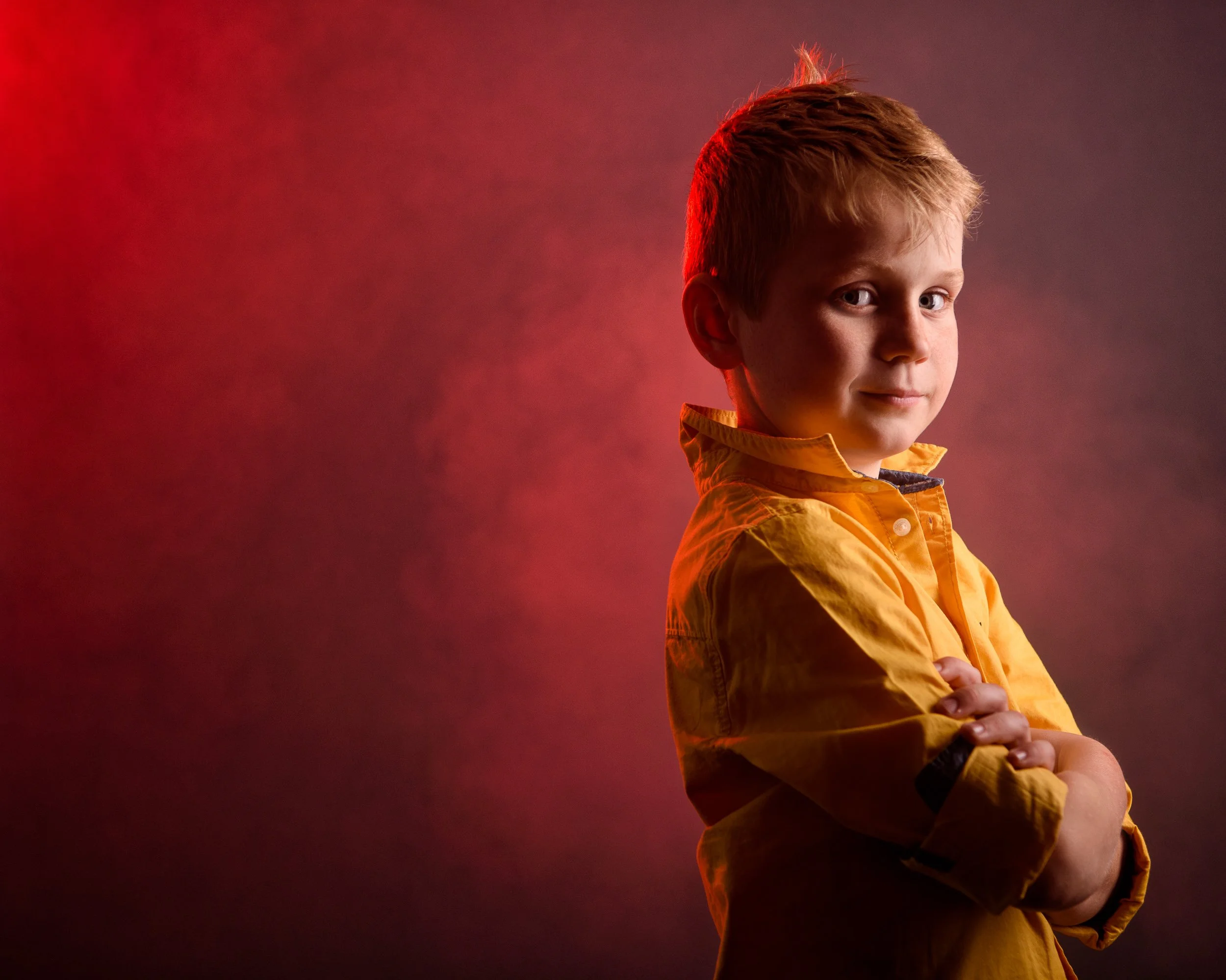A young boy with blonde hair wearing a yellow jacket, standing with arms crossed, looking at the camera against a red and purple gradient background.