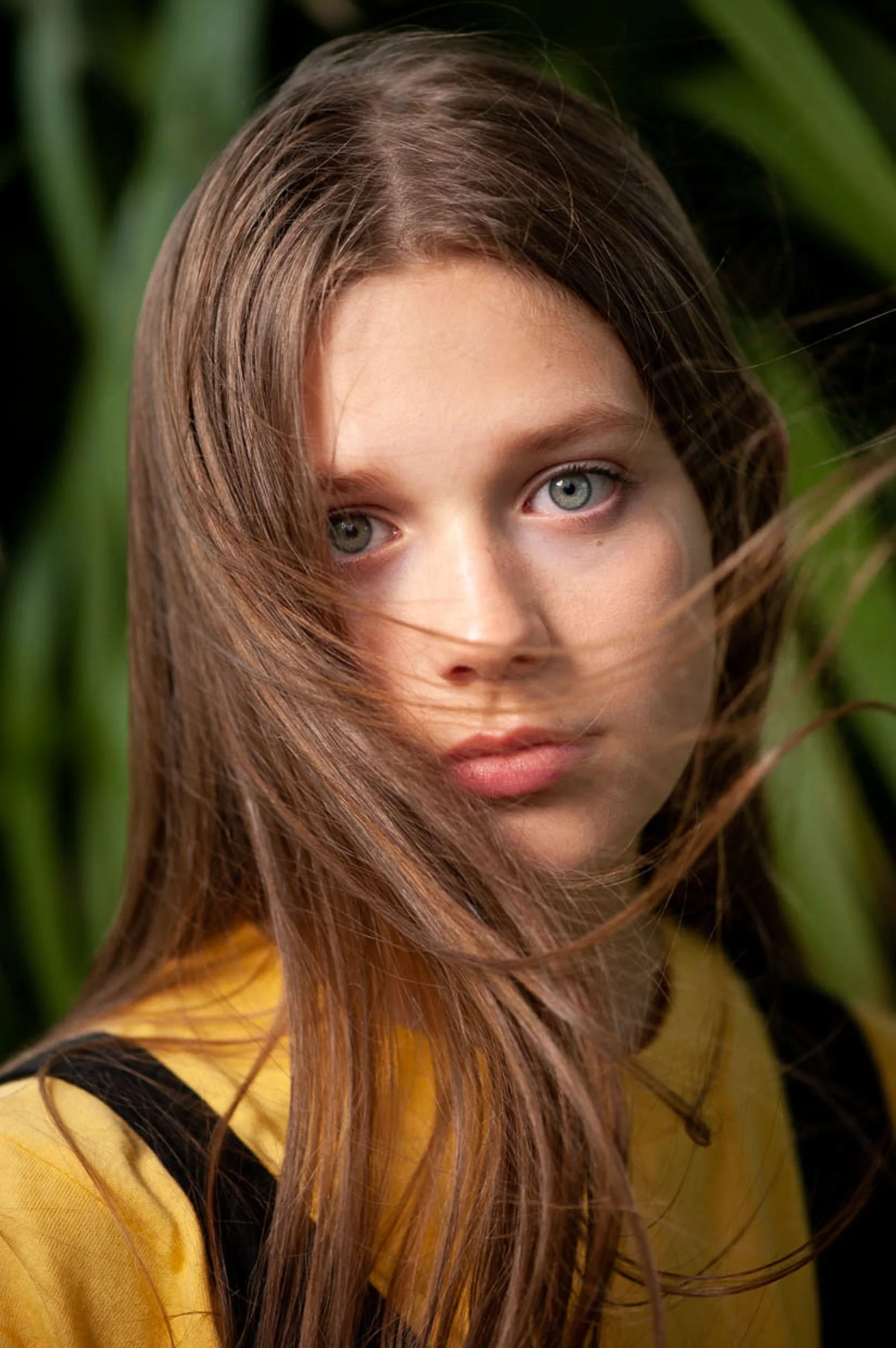 Close-up portrait of a young woman with long brown hair and blue eyes in natural light with greenery
