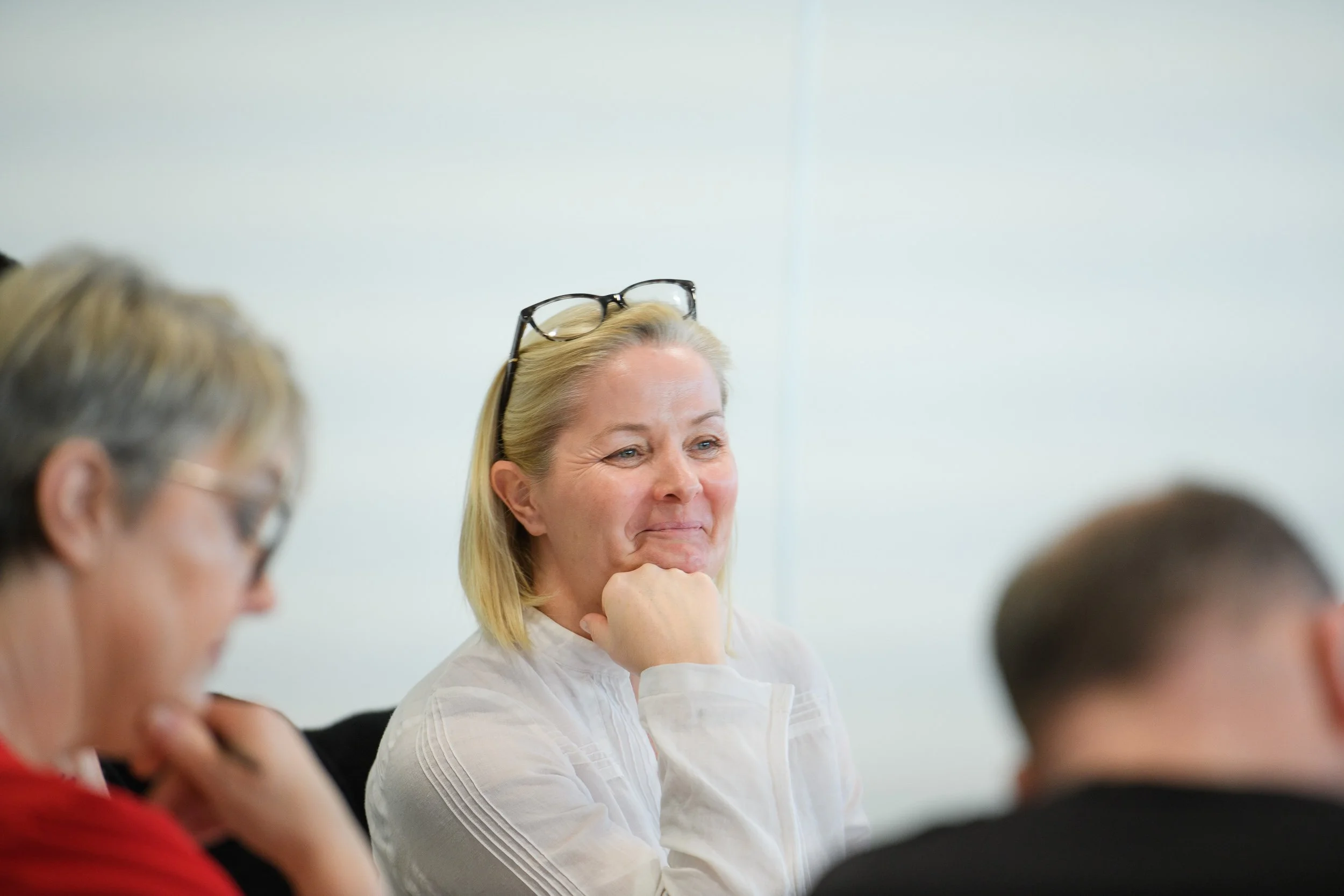A woman with blonde hair and glasses on her head, smiling with her hand resting on her chin, sitting at a table with two other people during a meeting or discussion.