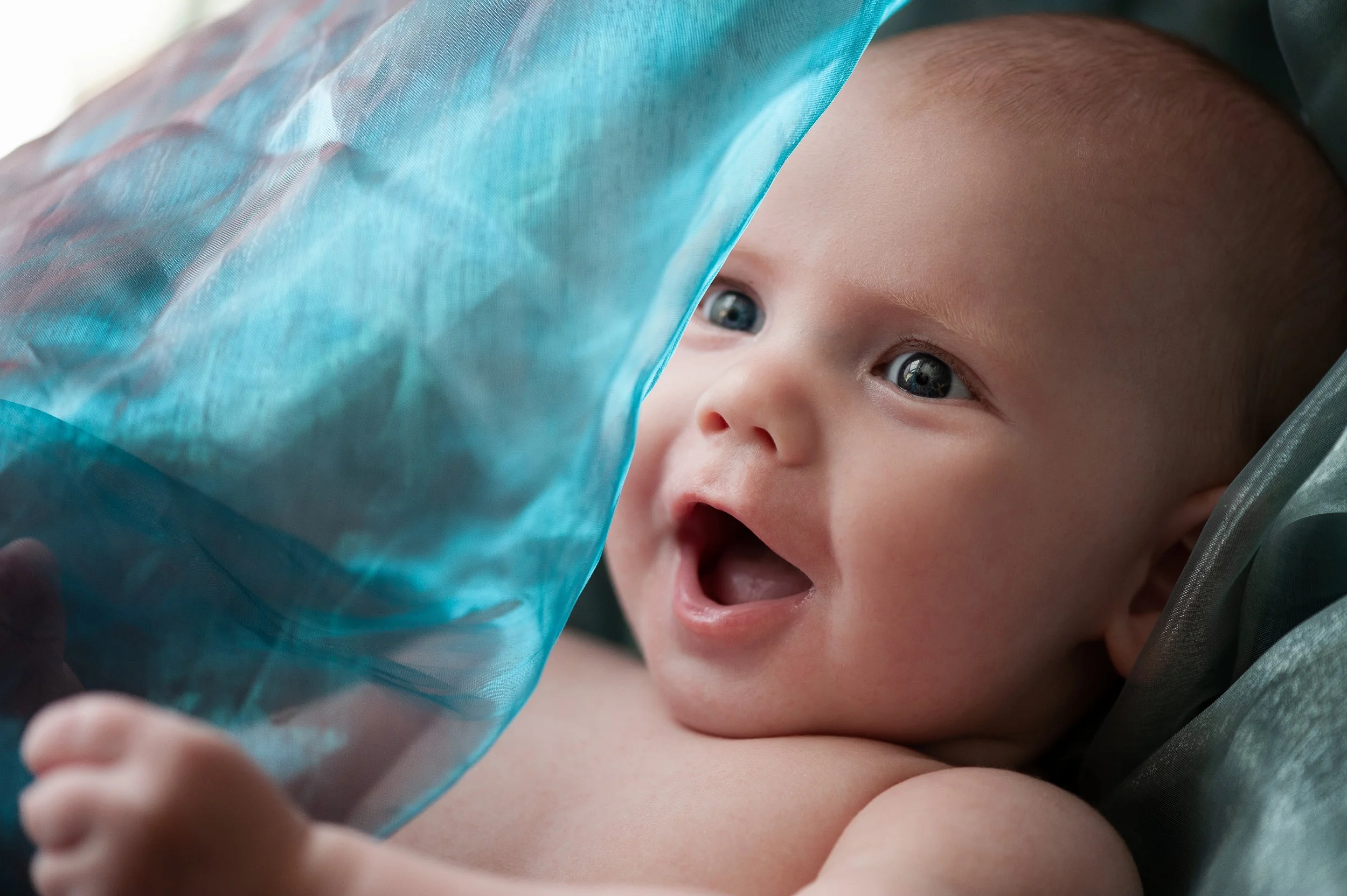 Close-up of a baby with blue eyes smiling and lying on a surface, partially covered by a blue translucent fabric.