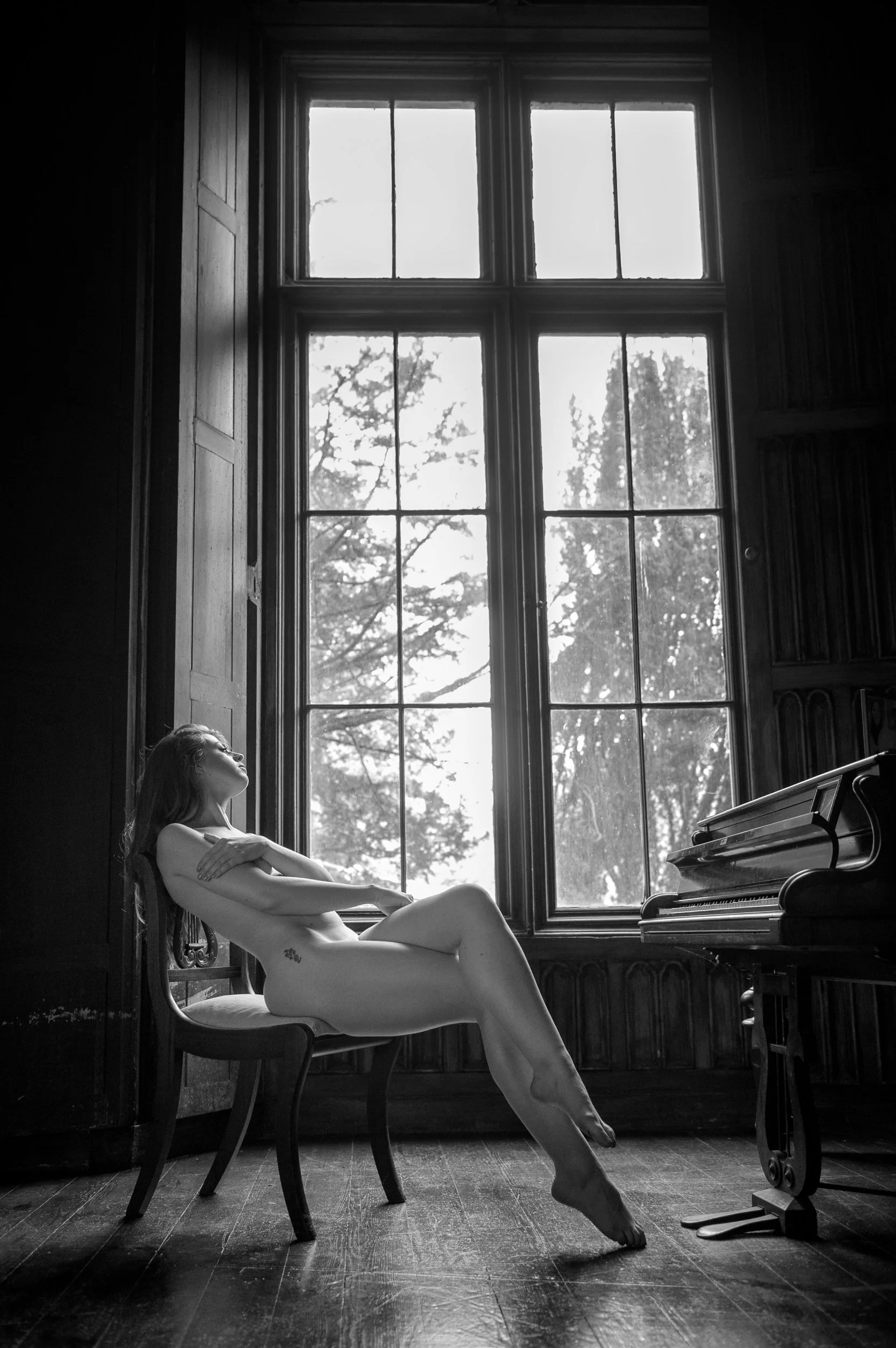Black and white portrait of a woman seated on a chair beside a grand piano, framed by large windows and wood-panelled interiors of Charleville Castle Tullamore
