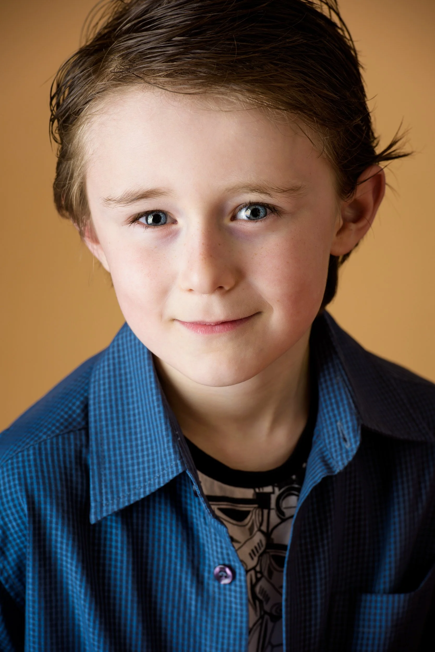 Close-up portrait of a young boy with light skin, blue eyes, and brown hair styled to the side, wearing a dark blue checkered shirt and a graphic t-shirt underneath, against a warm beige background.
