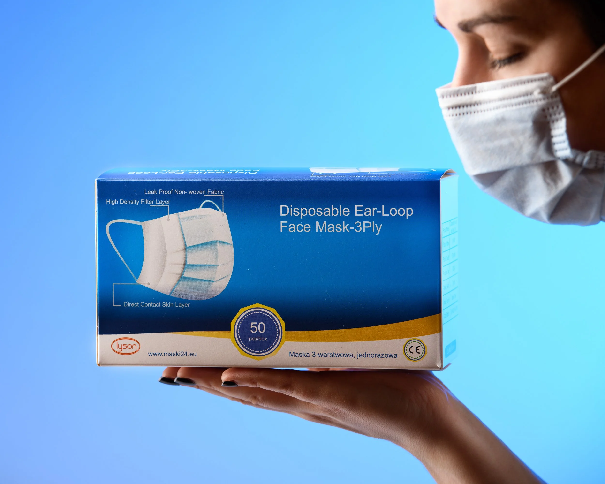 Person wearing a face mask holding a box of disposable masks against a blue background.
