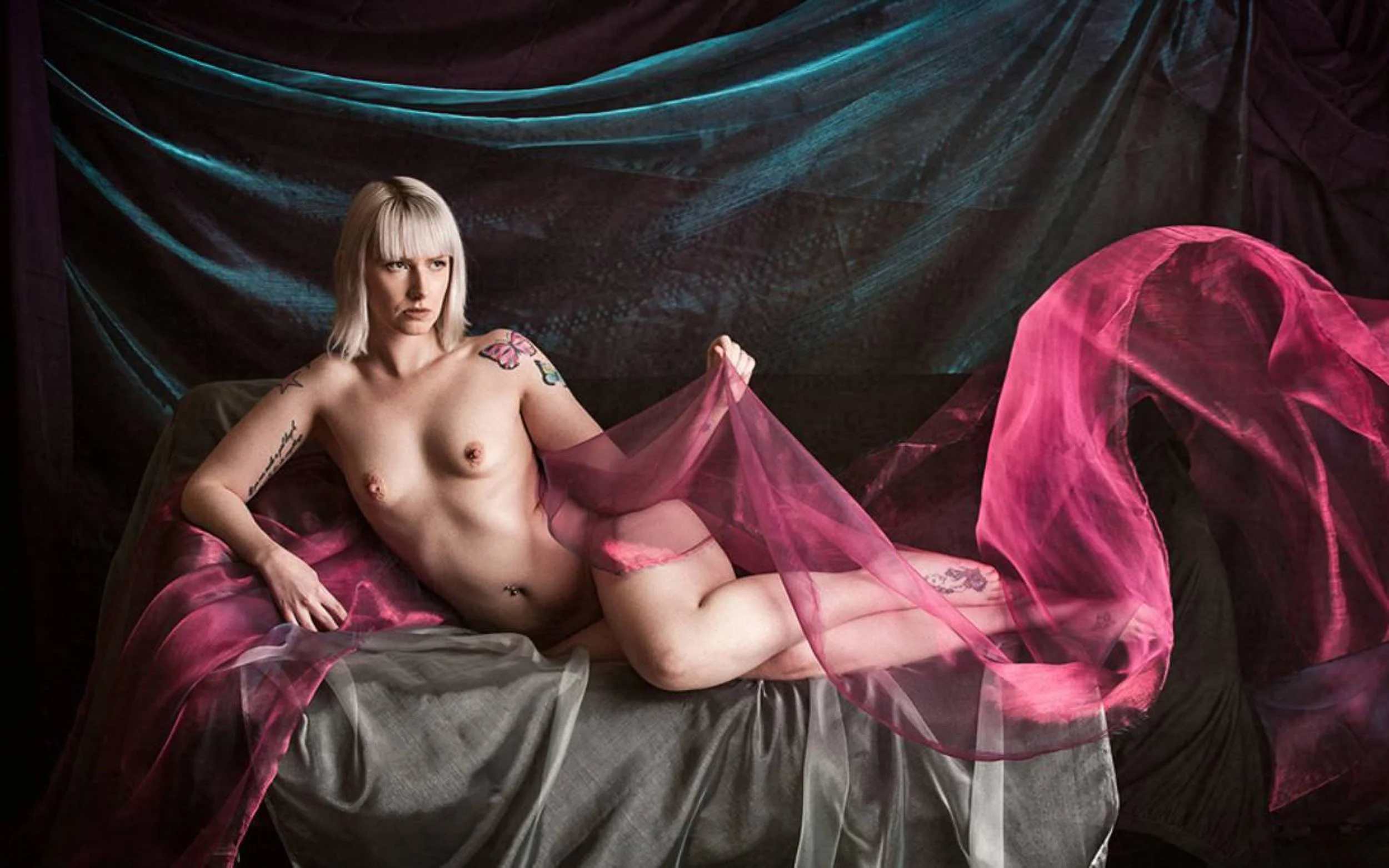 A woman with blonde hair and tattoos reclining on layered fabrics, set against a textured dark backdrop with soft coloured accents.

