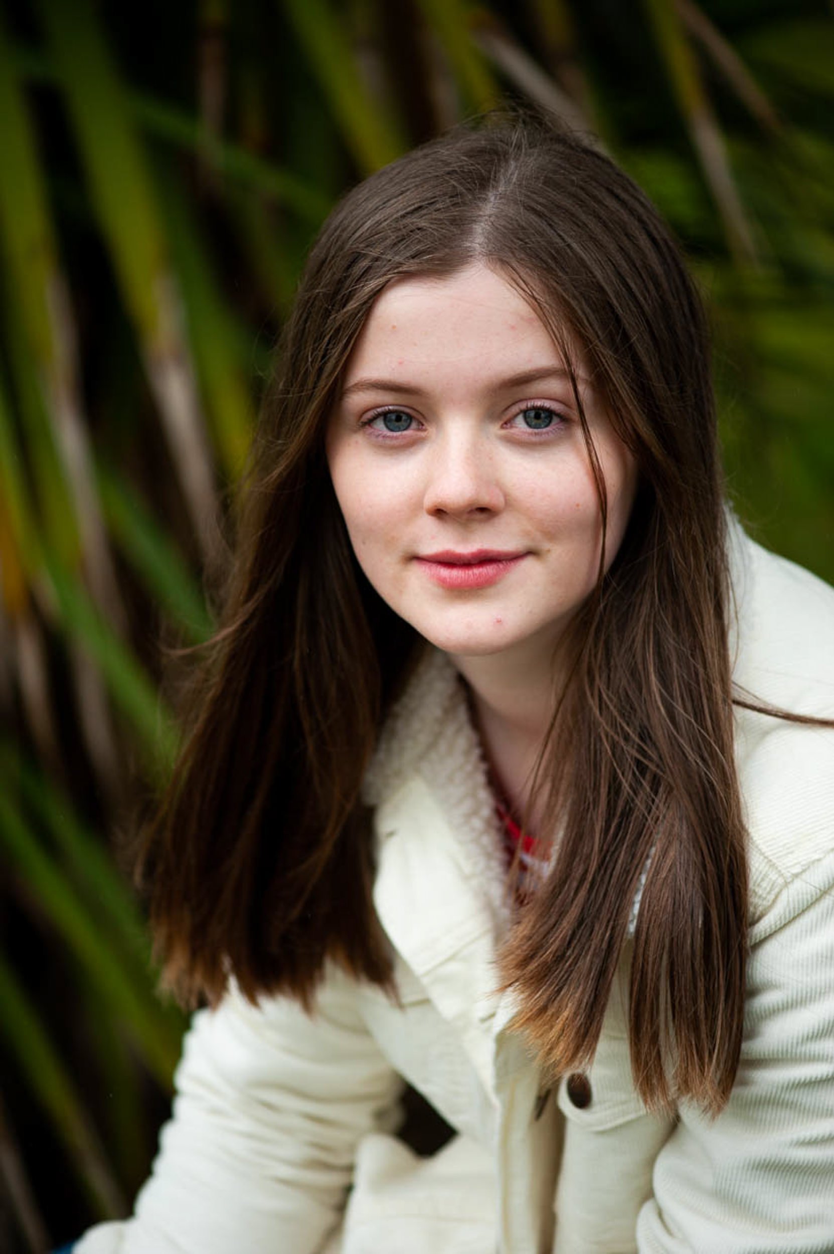 Outdoor portrait of a young woman with long brown hair and blue eyes surrounded by soft green foliage
