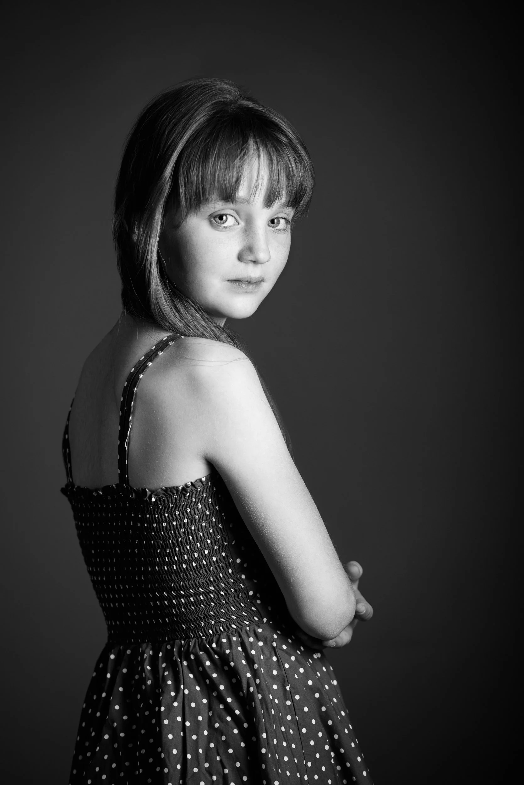Black and white portrait of a young girl with straight hair and bangs, wearing a polka dot dress, looking over her shoulder at the camera.
