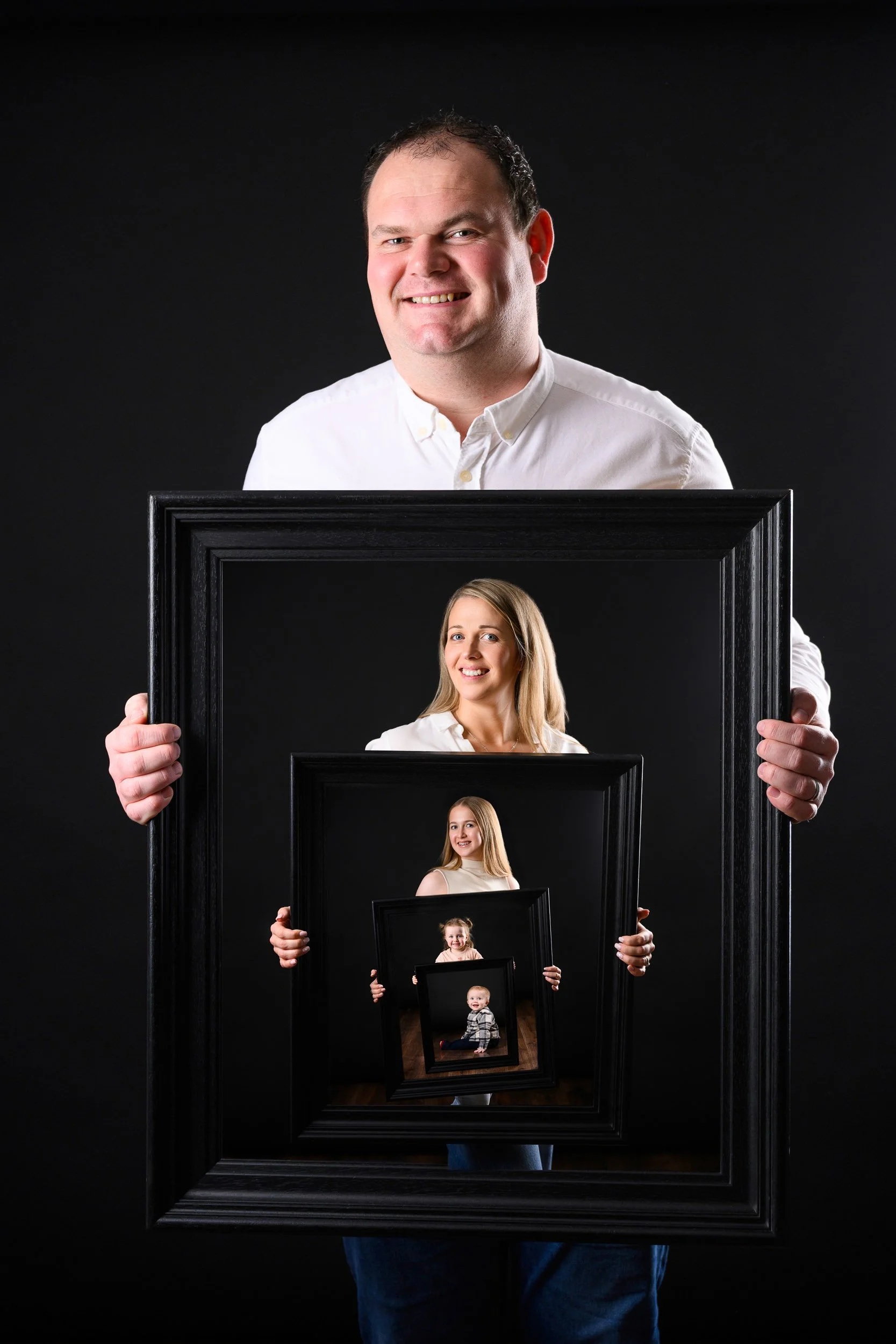 A man holding a picture frame with four nested portraits inside, set against a black background.
