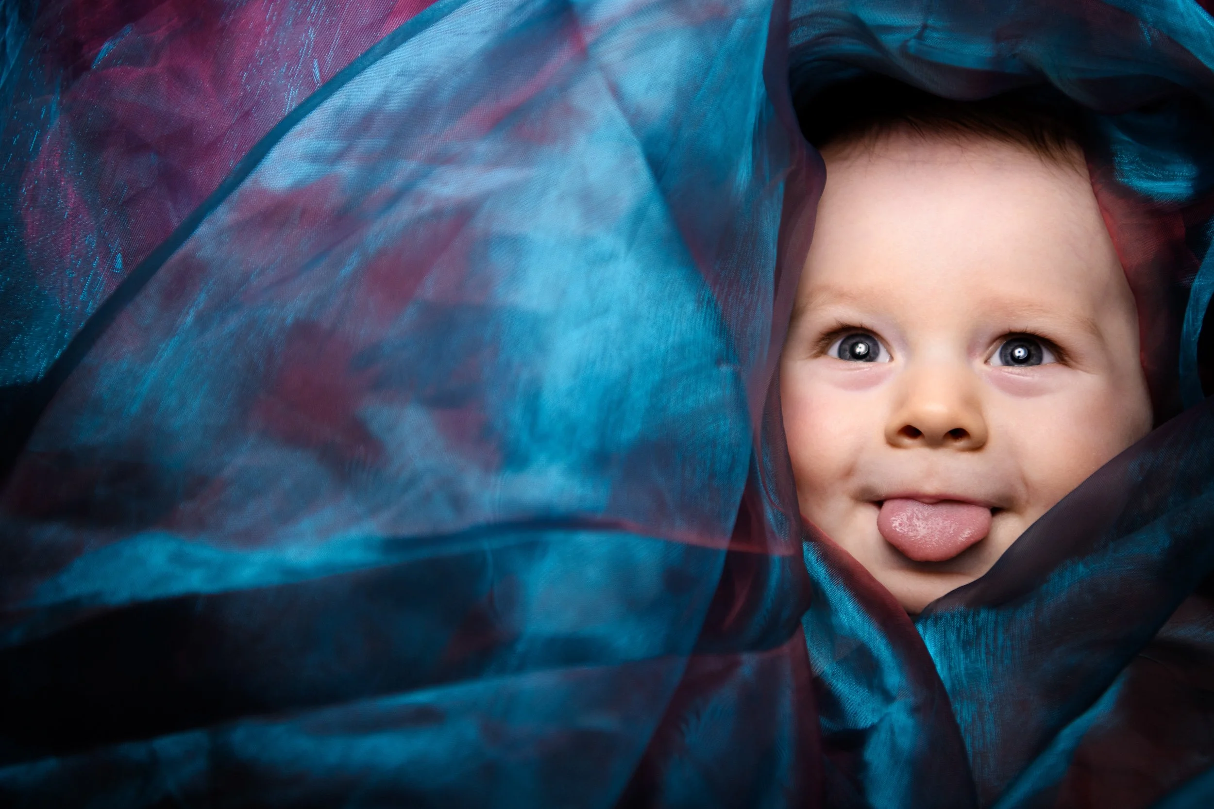 A baby with blue eyes and light brown hair playfully sticking out their tongue under sheer colourful fabric.
