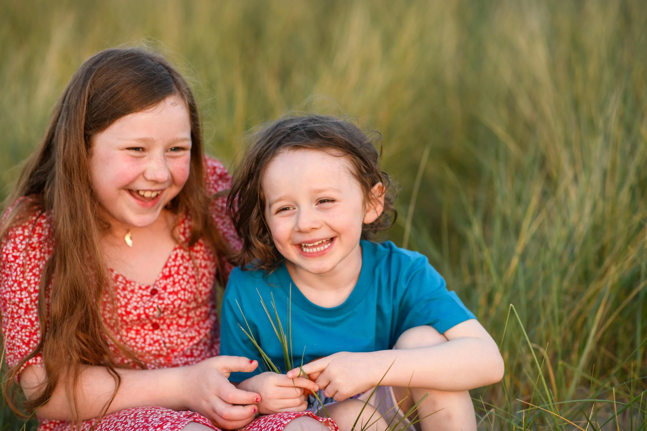Two children, a girl and a boy, sitting on grass in a field, smiling and enjoying each other's company.